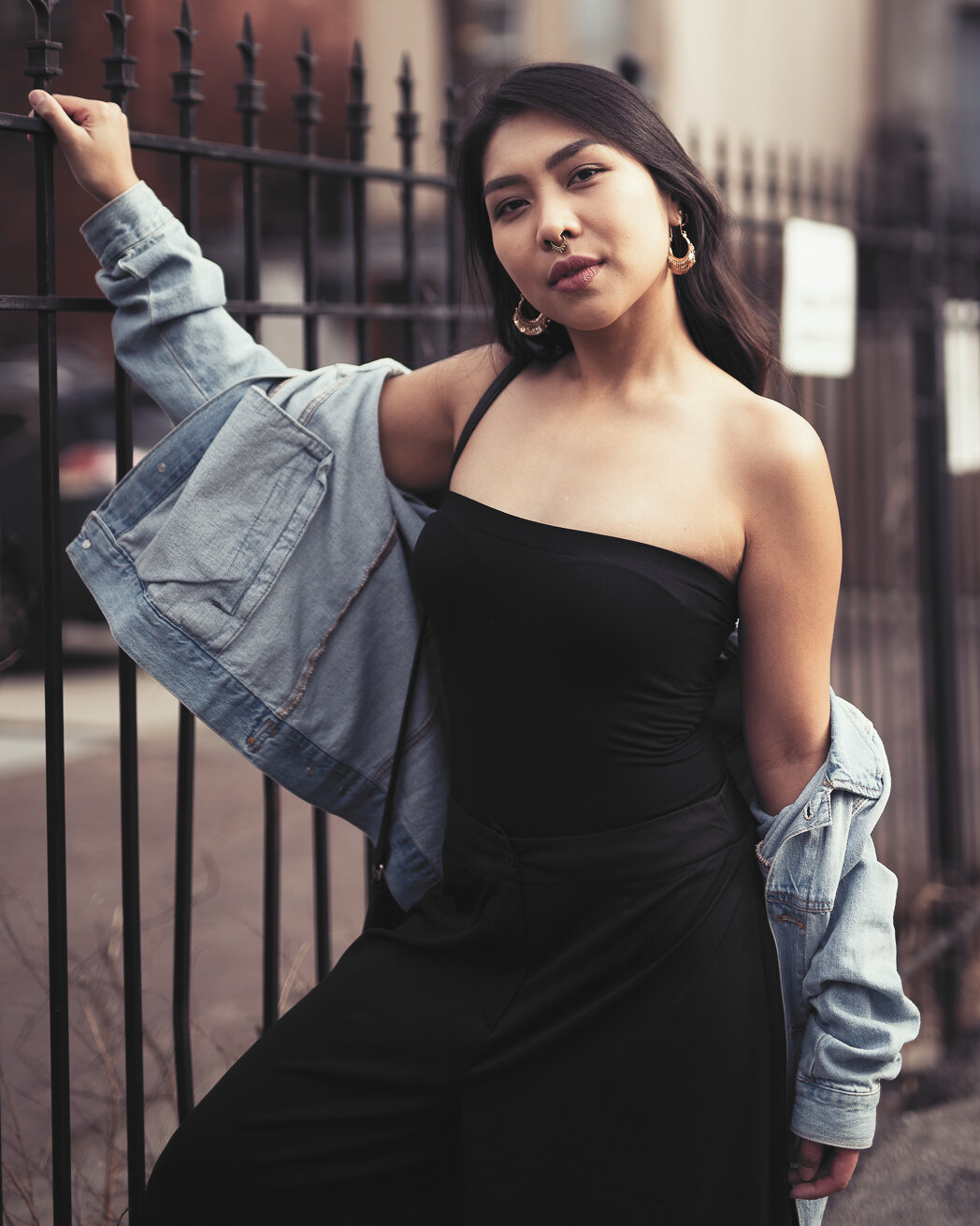 A confident young woman with shoulder-length dark hair standing outdoors near a black metal fence in an urban area. She is wearing a black strapless top, black pants, and a slightly oversized denim jacket draped off her shoulder. She has hoop earring