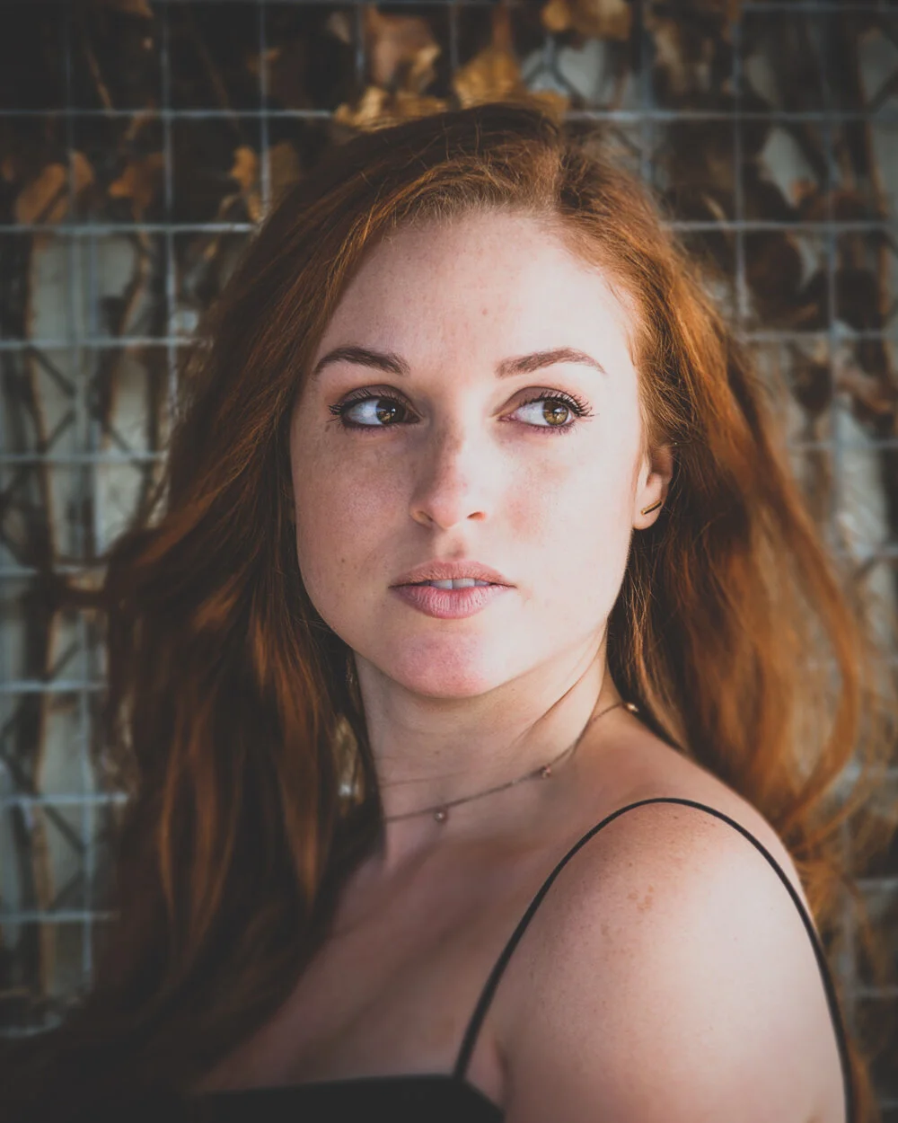 Close-up of a young woman with red hair and freckles looking sideways, with a blurred background of a wire fence with some leaves.
