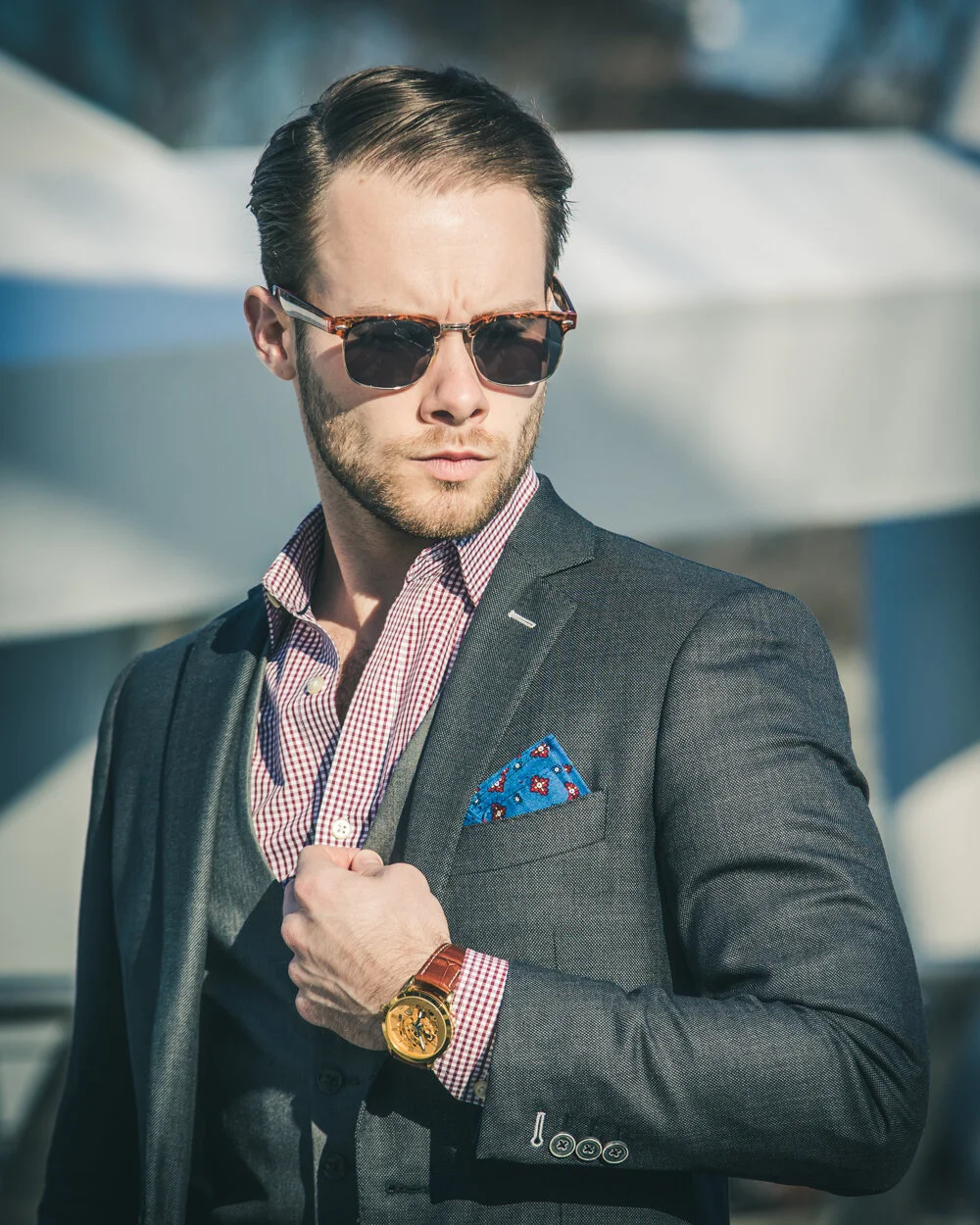 A man in a suit and sunglasses outdoors, adjusting his jacket and looking away.