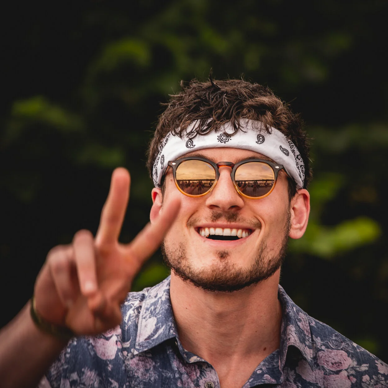 A young man smiling and making a peace sign with his right hand outdoors, wearing sunglasses, a white bandana, and a floral shirt.