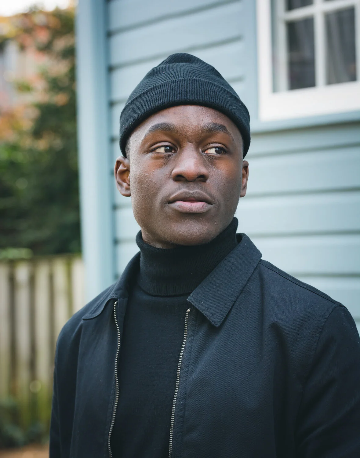 A young man wearing a black beanie and black jacket standing outside near a house with siding, looking off to the side.