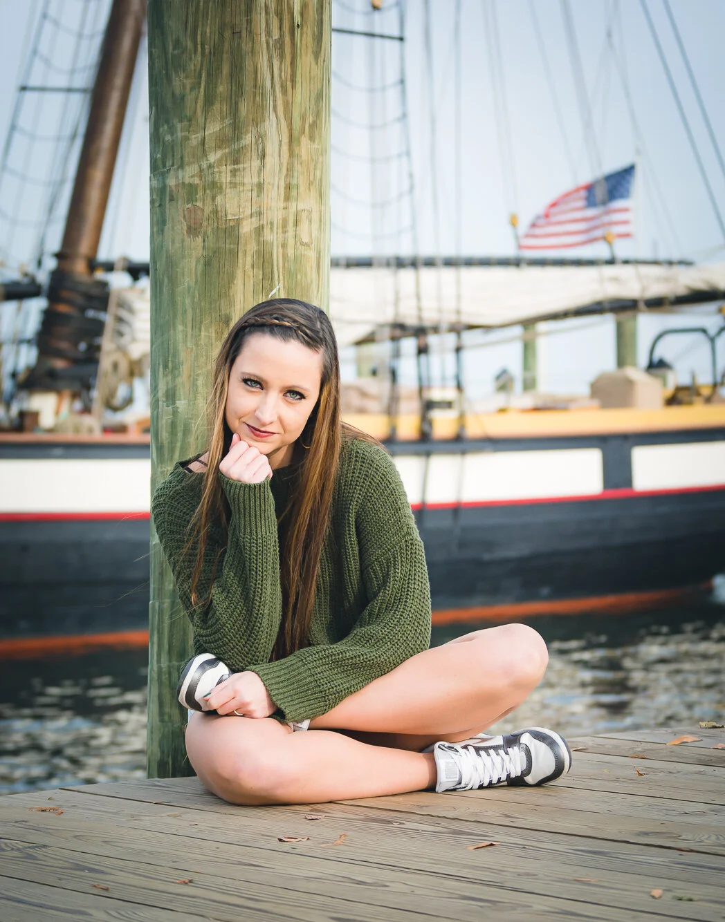 A young woman sitting on a wooden dock with a large boat and an American flag in the background.
