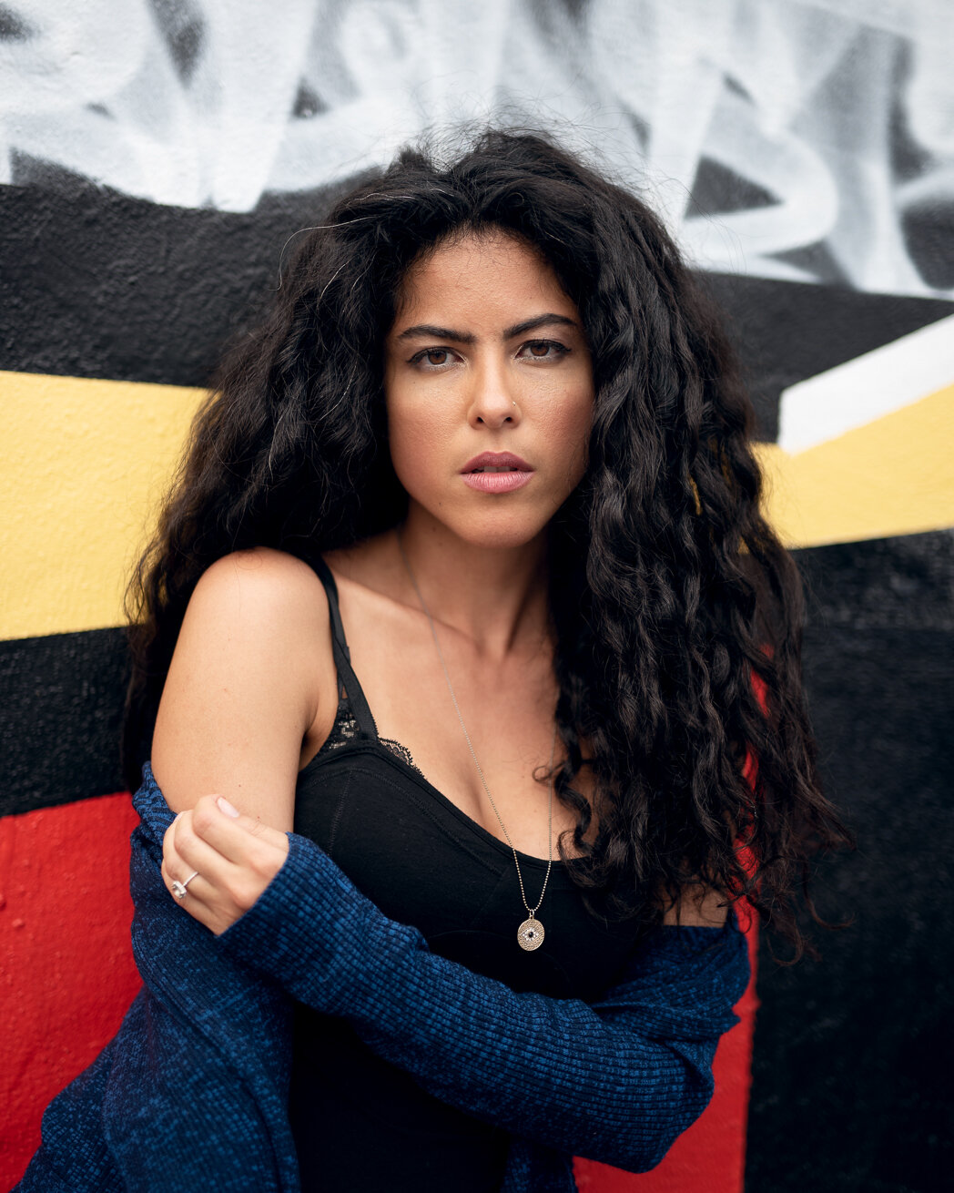 A woman with long, curly black hair wearing a black camisole and a blue cardigan, standing against a graffiti wall with black, yellow, white, and red colors.