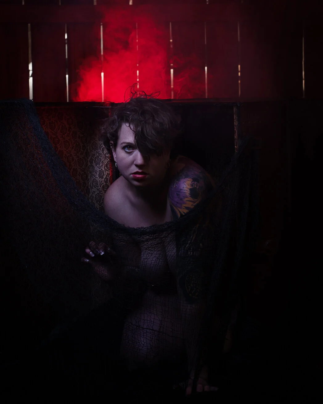A woman with short curly hair and tattoos on her shoulder appears to be partially nude, crouching behind a black lace fabric in a dimly lit room with red lighting and wooden panels in the background.