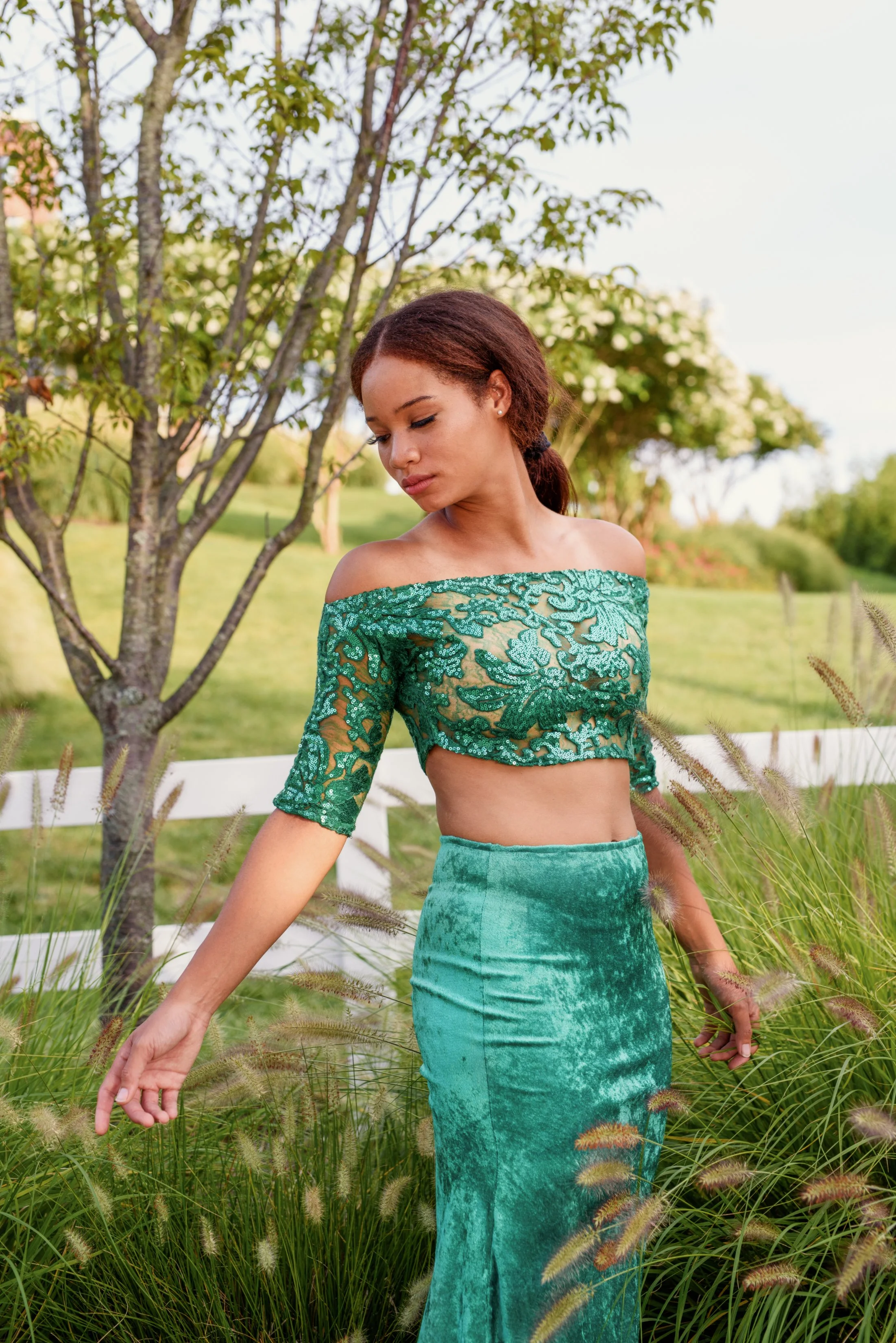 A young woman with curly brown hair in a ponytail, wearing an off-the-shoulder green lace crop top and high-waisted velvet skirt, stands in a grassy outdoor setting with trees and white fencing in the background.