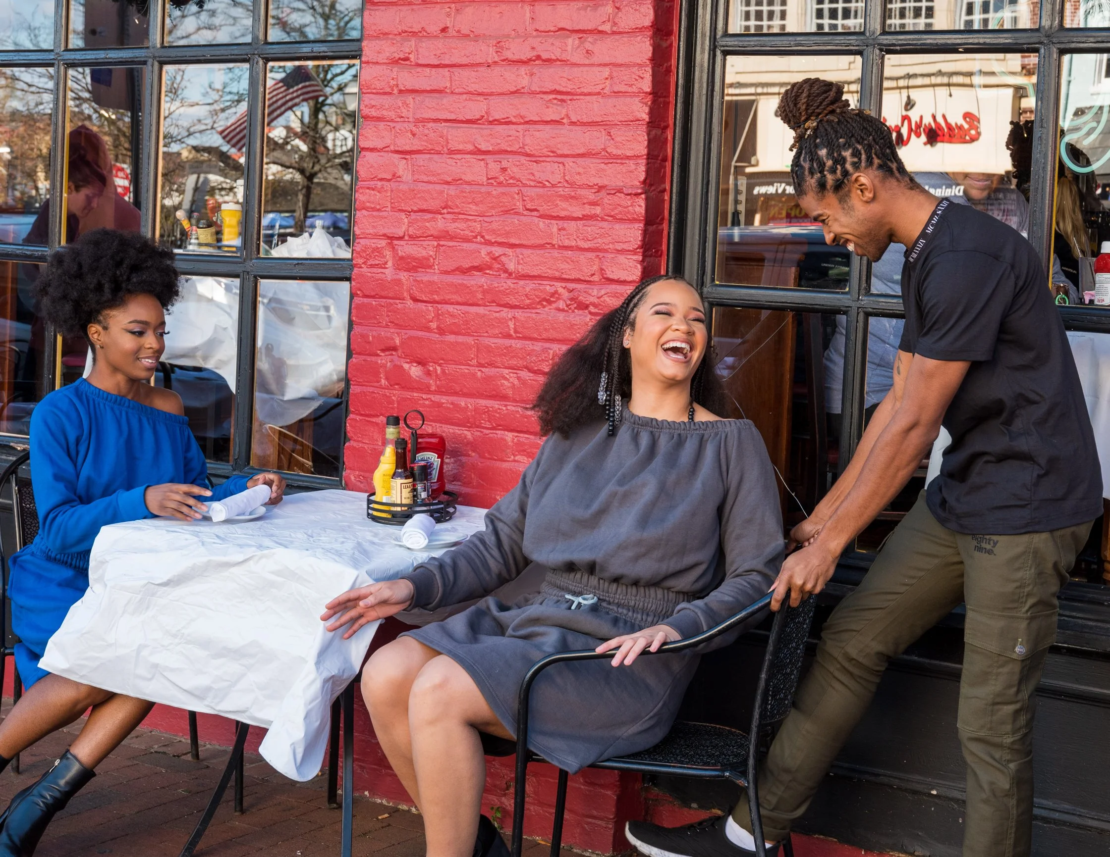 Three women are outside a restaurant, with two of them sitting at a table and one standing. The seated woman is laughing and the standing woman is smiling. The third woman is sitting at a table nearby, looking at the others. There are condiments on t