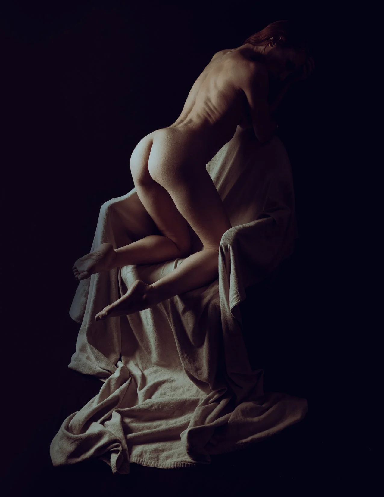 A nude woman sitting on a draped cloth, leaning forward, with her hands on her head and face partially obscured in shadow.