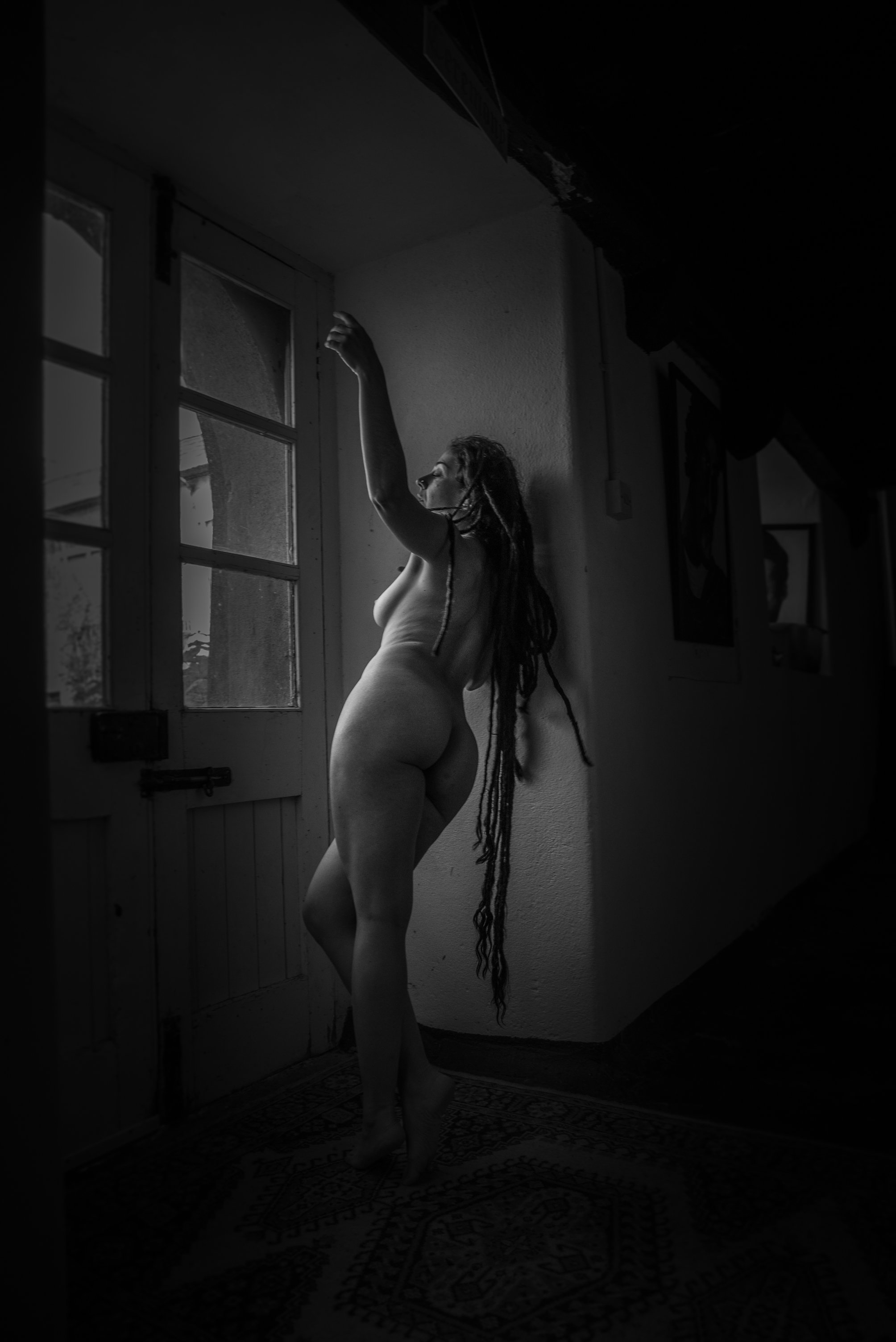 Black and white photo of a nude woman with long dreadlocks standing by a window, looking outside, with her arm raised and one leg crossed over the other.