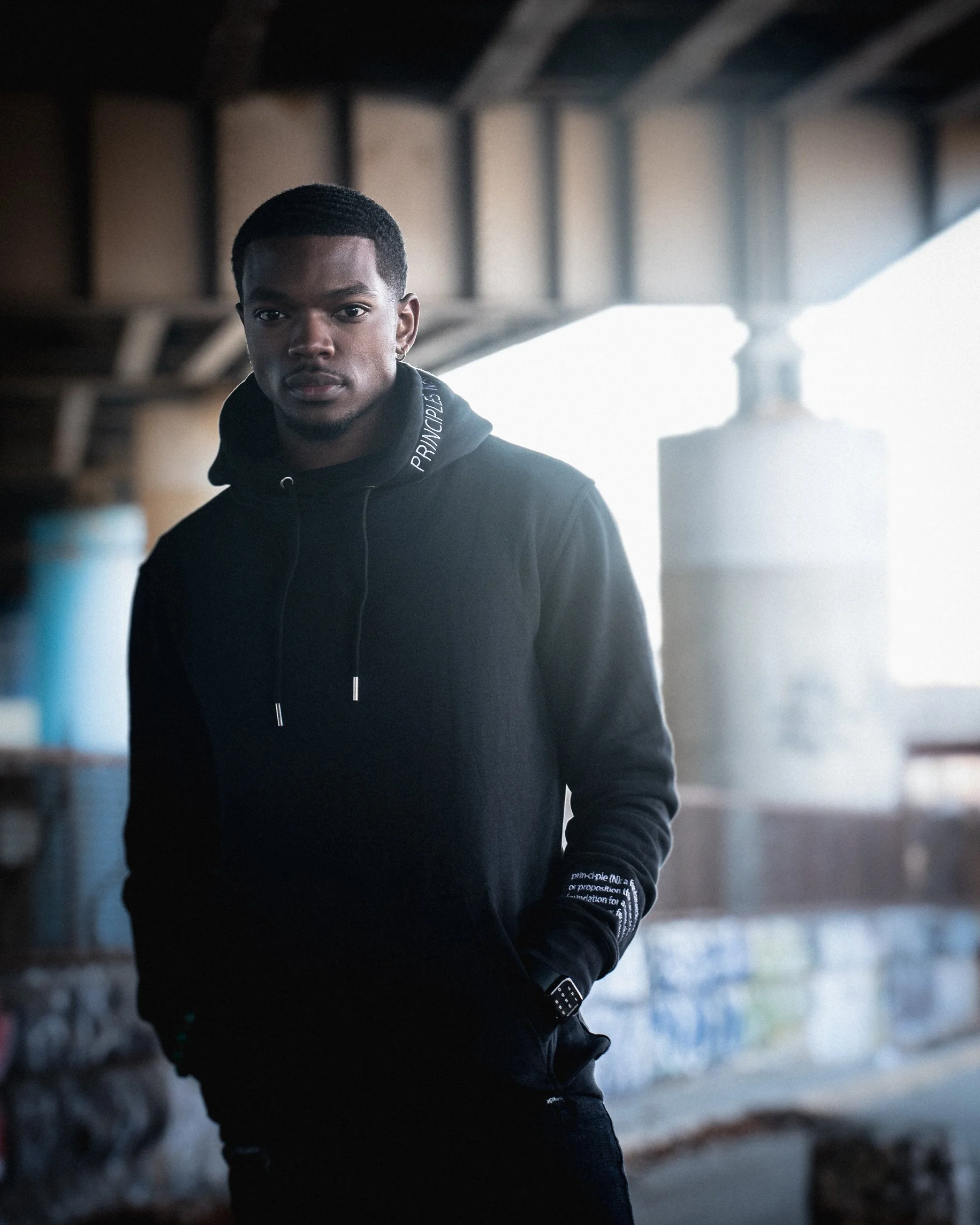A young man standing under a bridge, wearing a black hoodie with text and a pair of gloves, with urban graffiti and structures in the background.