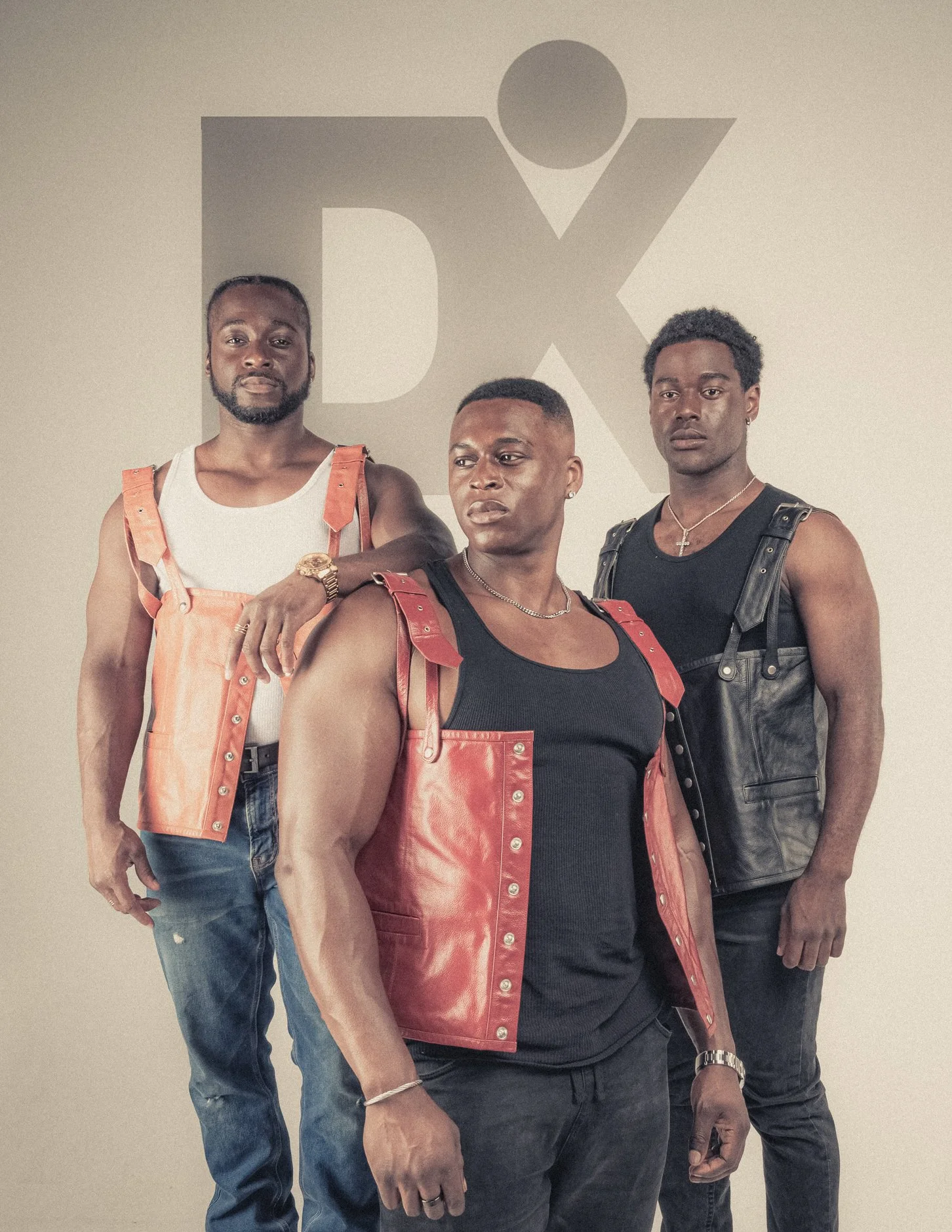 Three men standing in front of a wall with a DX logo. They are wearing sleeveless black shirts with orange and black leather vests, posing confidently.
