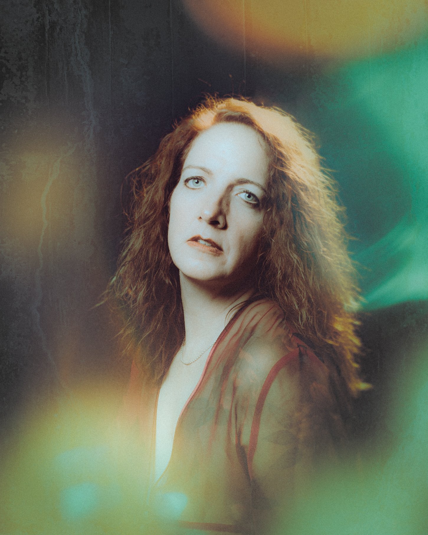 Portrait of a woman with curly red hair and blue eyes, wearing a sheer red top, against a dark background with multicolored light flares.
