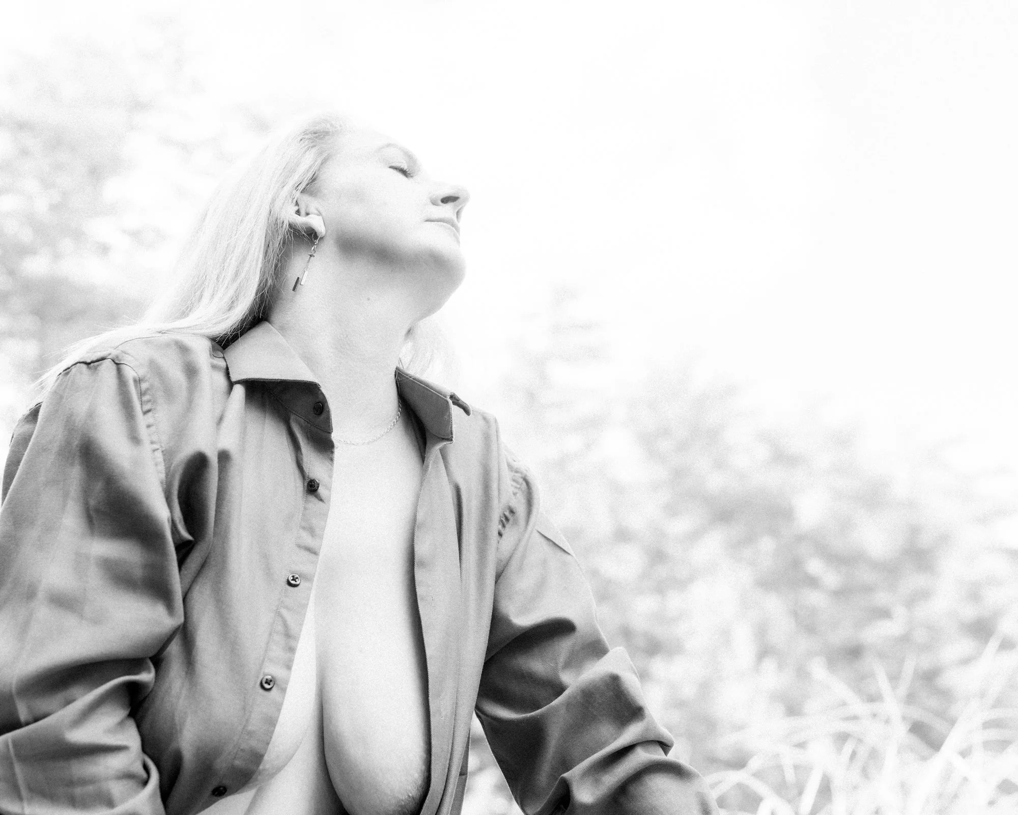 A woman with long hair with her eyes closed, wearing a button-up shirt and earrings, standing outdoors with a bright sky and blurred trees or foliage in the background.