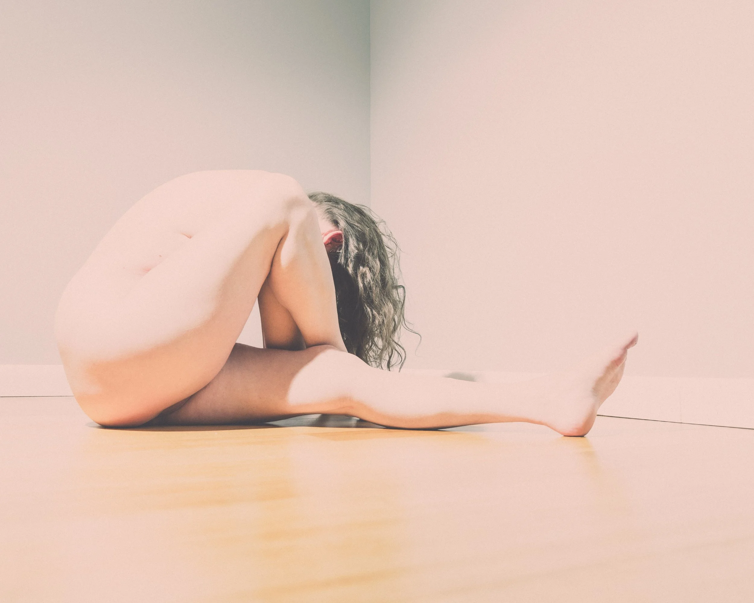 Person with long curly hair sitting on a wooden floor in a corner, hugging their knees with their head resting on their arms.