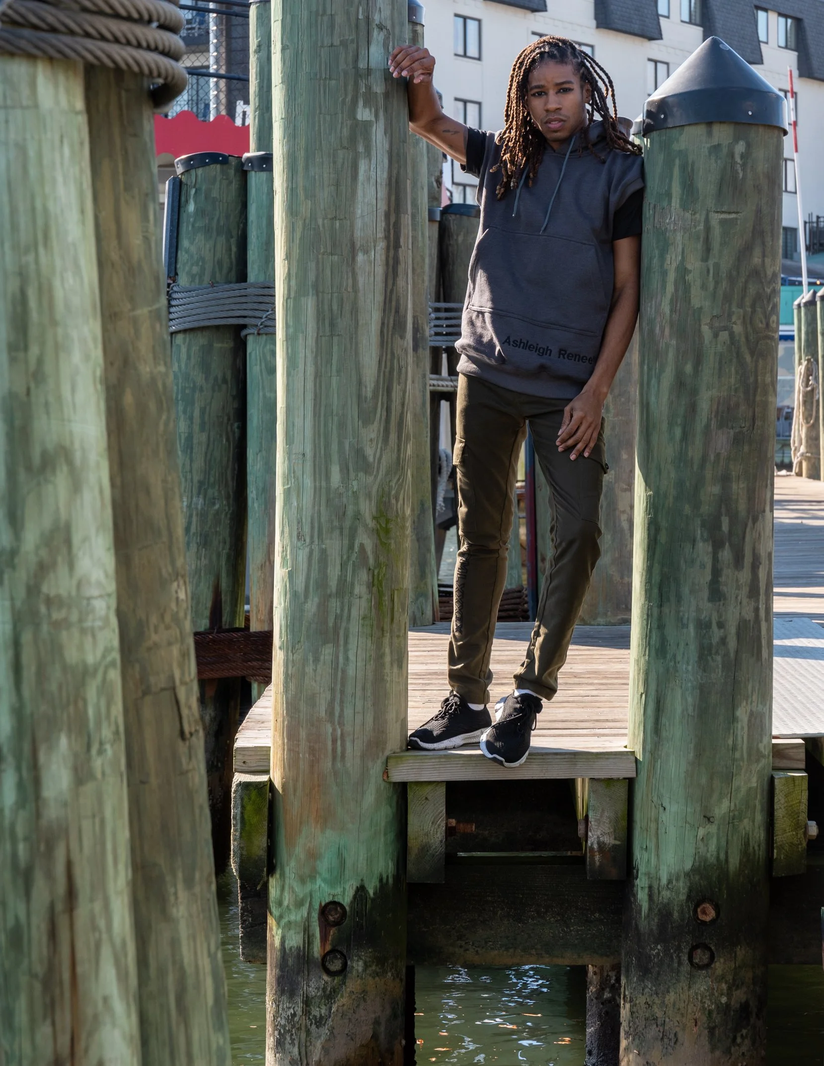 A young man with dreadlocks standing on a small wooden platform attached to a pier, holding onto a large wooden post, in a waterfront area with buildings in the background.