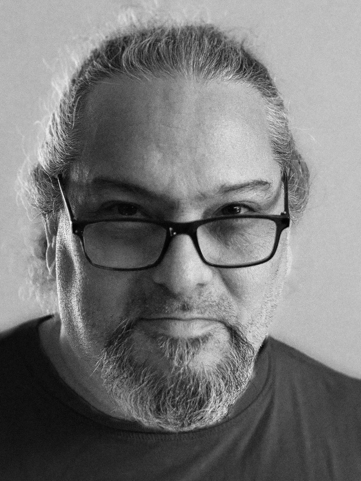 Black-and-white close-up photo of a middle-aged man with long hair tied back, glasses, and a beard, looking at the camera.