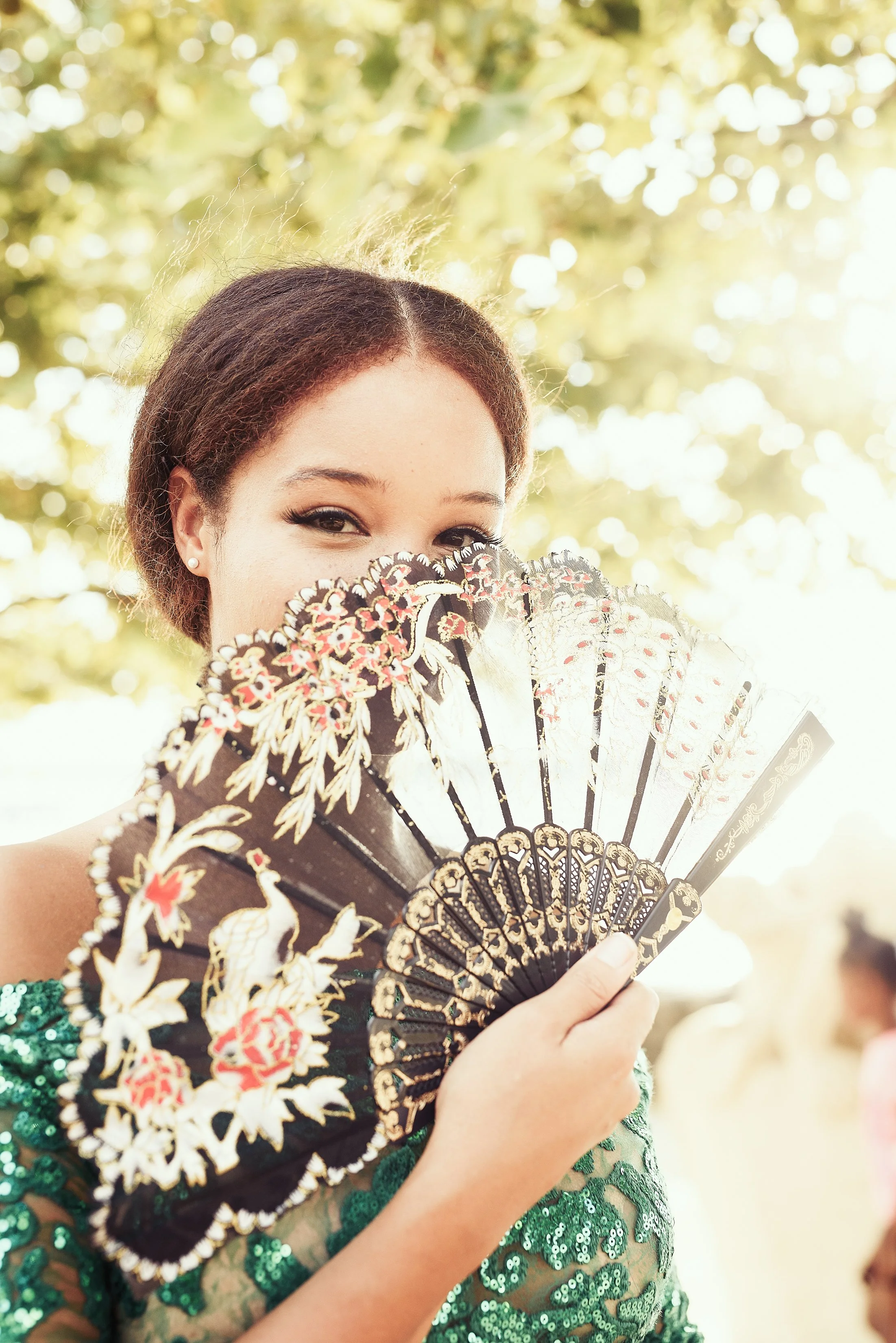 A woman holding an ornate hand fan partially covering her face, outdoors with sunlight filtering through trees in the background.