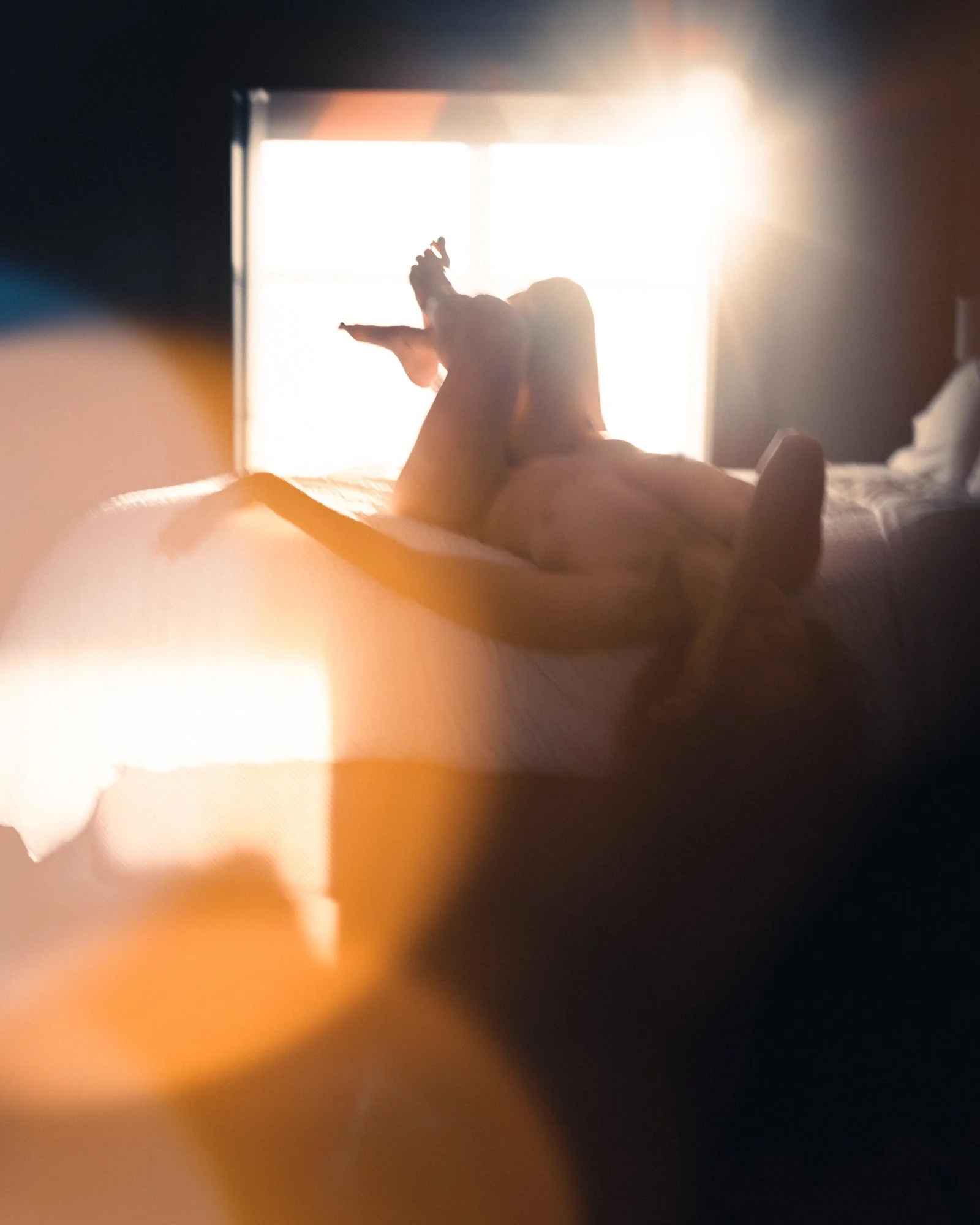 A person lying on a bed, back facing the camera, stretching or relaxing with arms raised, illuminated by bright sunlight coming through a window.
