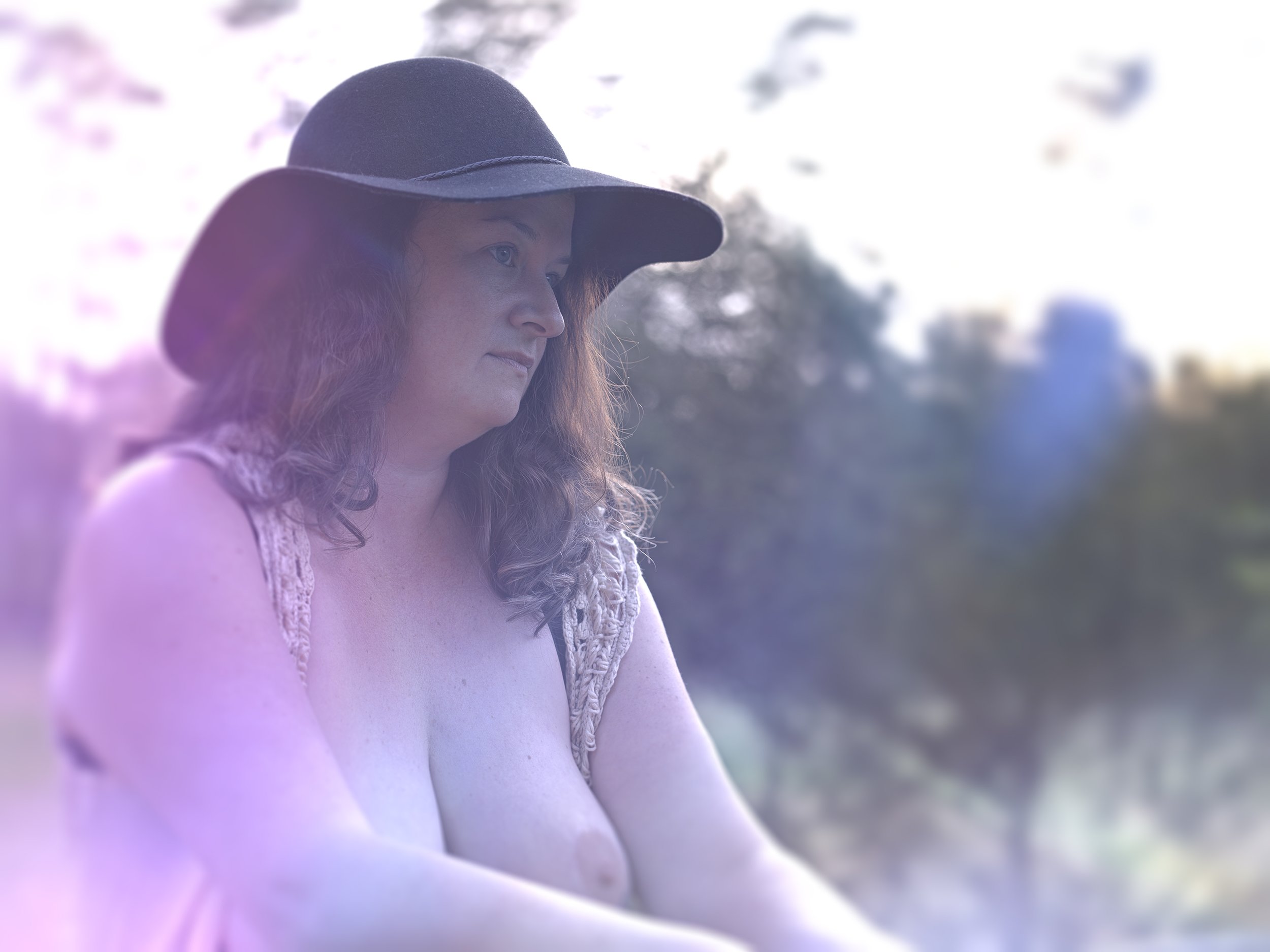 A woman wearing a large black hat, sitting outdoors with blurred trees and sky in the background, illuminated by sunlight.