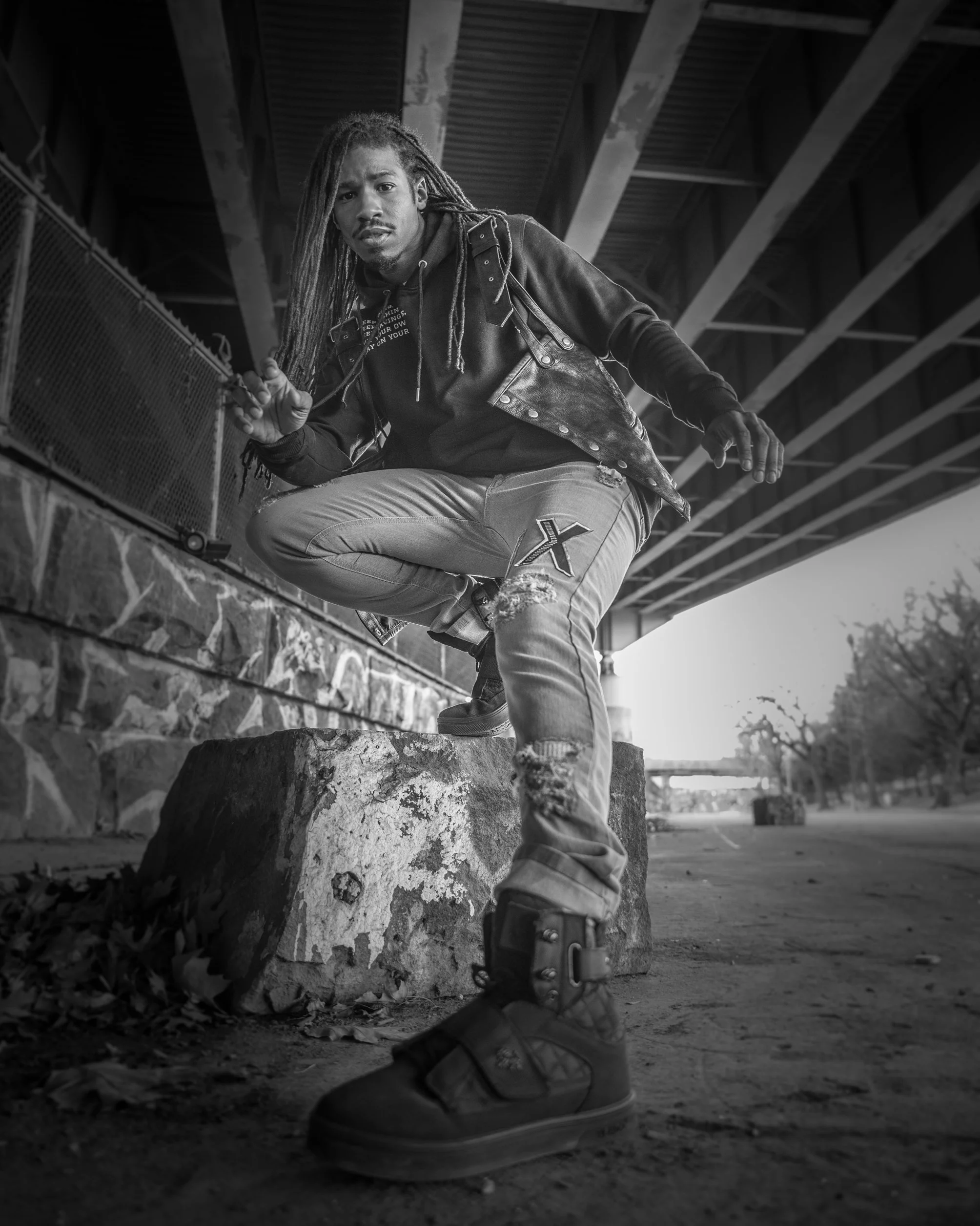 A man with dreadlocks crouches on a large rock under a bridge, looking at the camera. The photo is in black and white, with an urban setting and trees in the background.