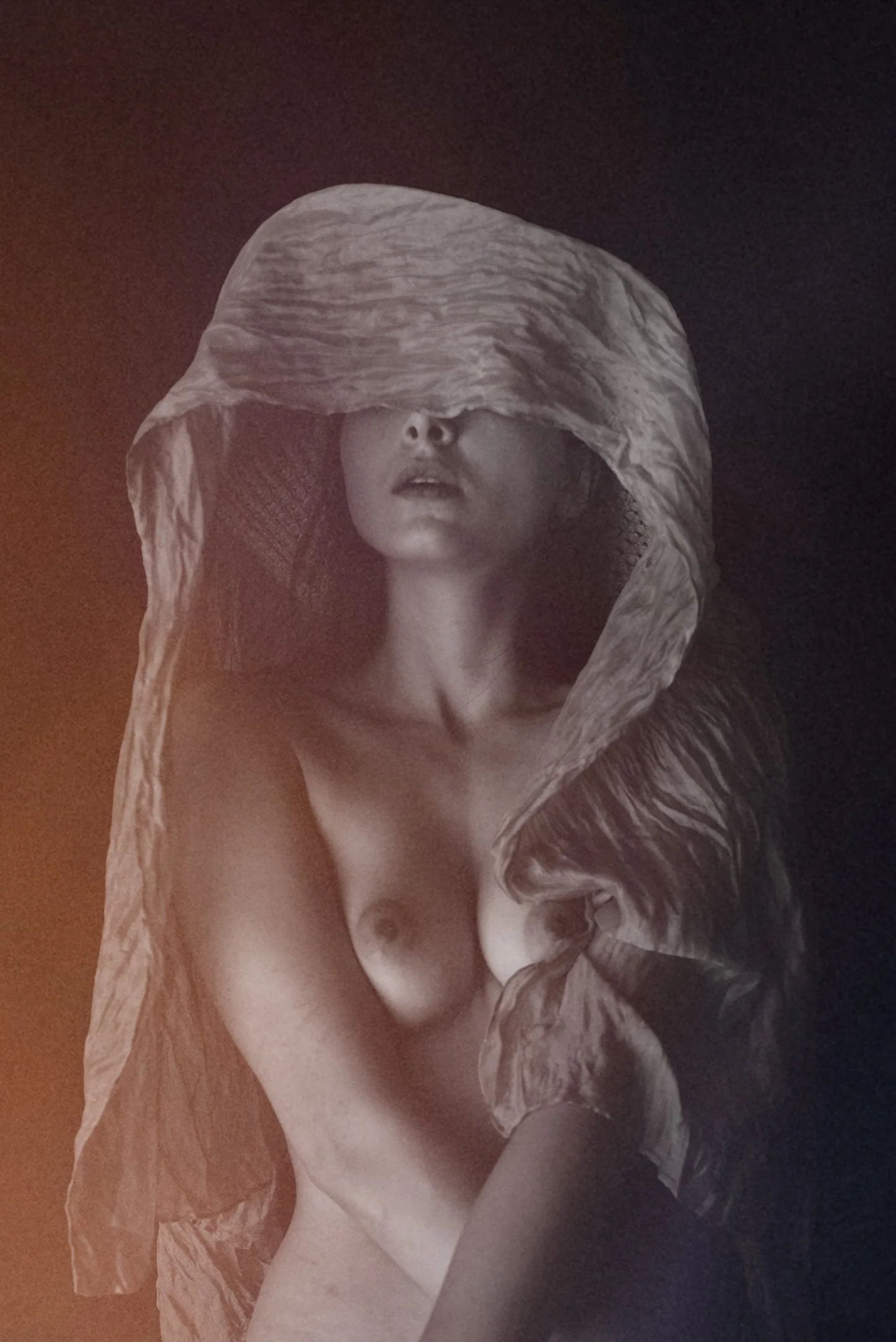 A black and white photo of a woman with her eyes covered by a large textured cloth, exposing her lips and upper chest, with soft lighting highlighting her face and body.