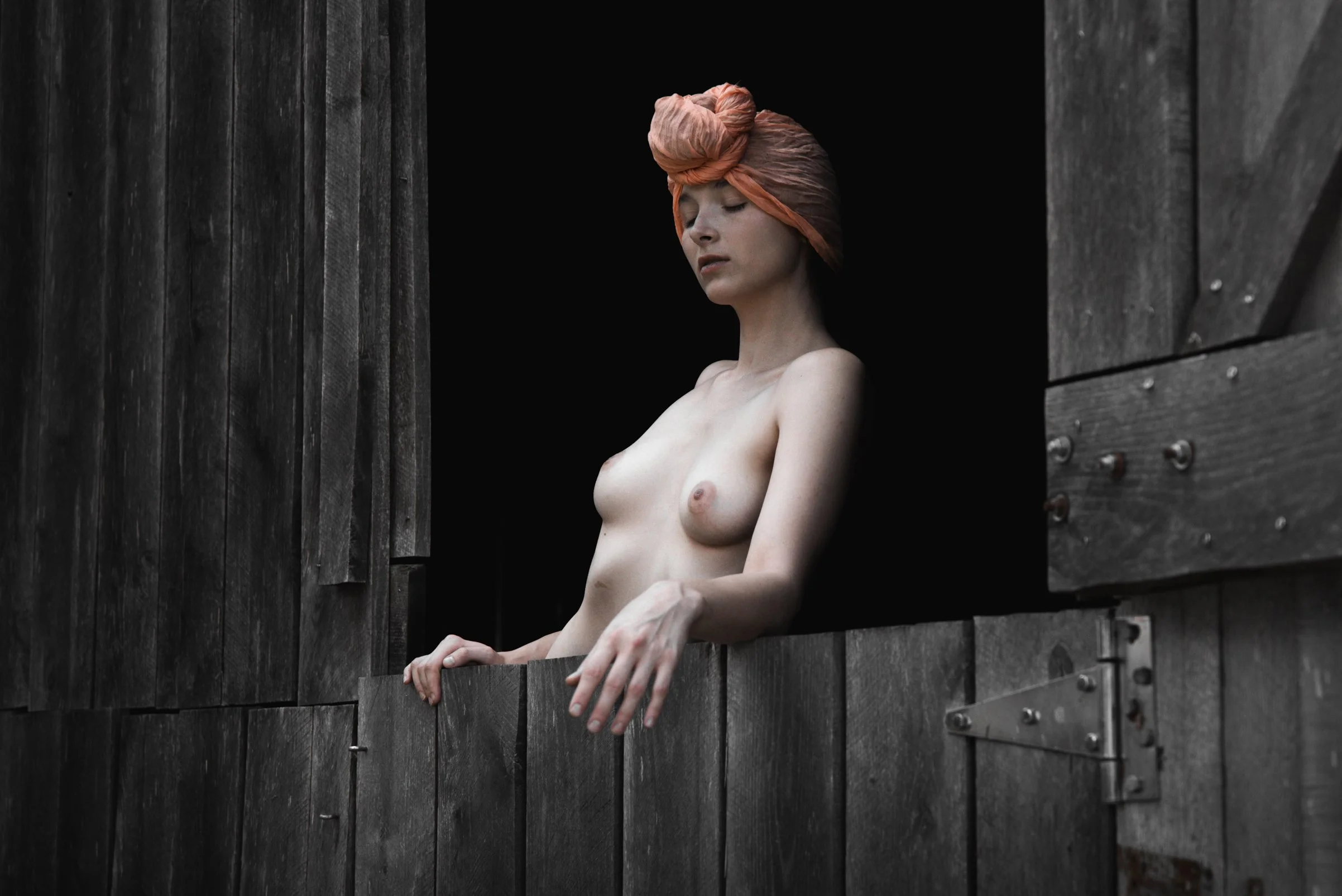 A topless woman with a pink headwrap standing at a wooden window, looking downward with her eyes closed, surrounded by dark wooden walls.