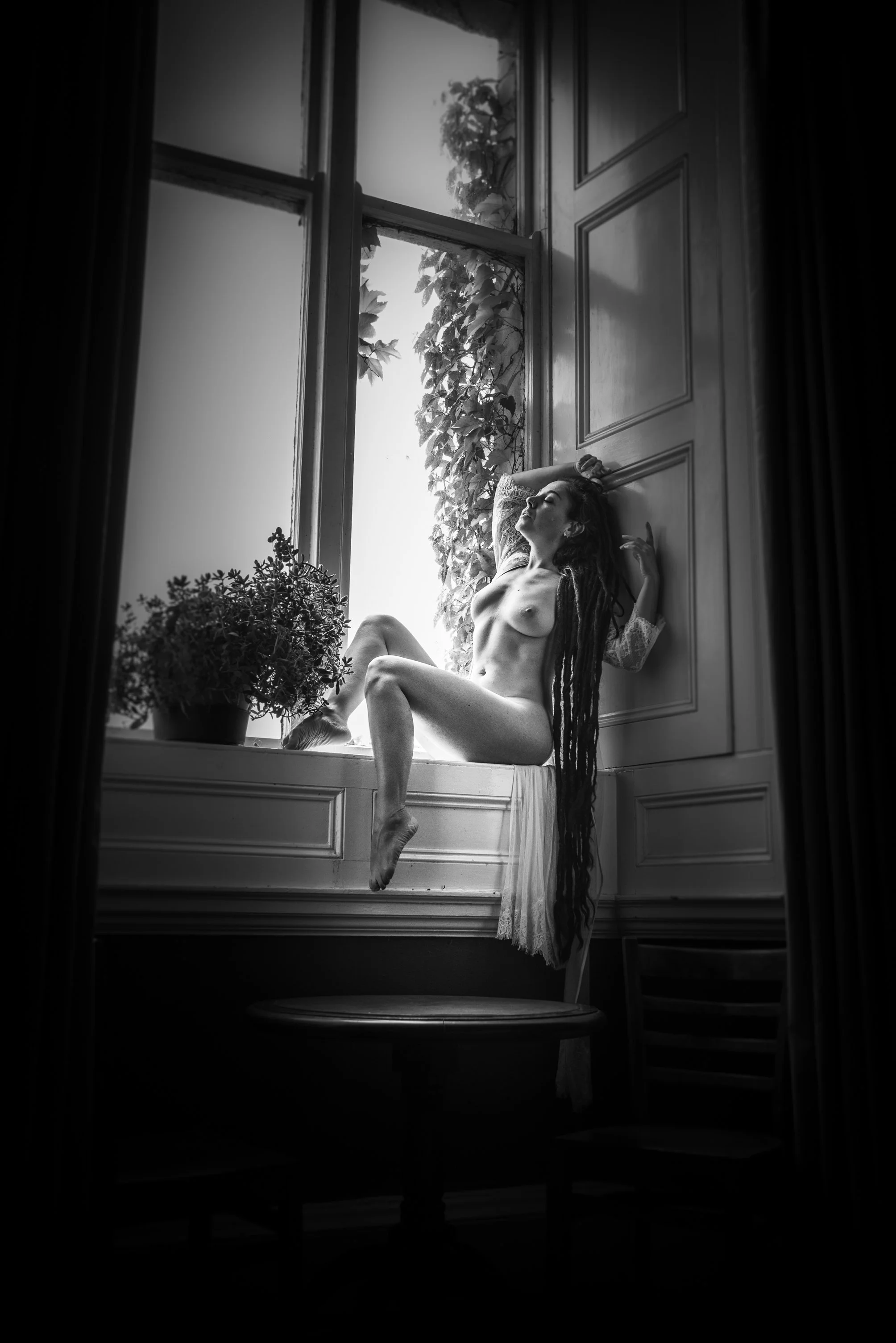 A black and white photo of a nude woman with long dreadlocks sitting on a window ledge, surrounded by plants, with sunlight coming through the window.