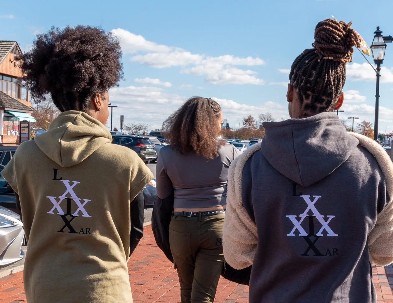 Three people walking outdoors on a sunny day, two wearing hoodies with a cross design on the back, and one person in the middle wearing a gray crop top and olive pants. There are cars parked and a building in the background.