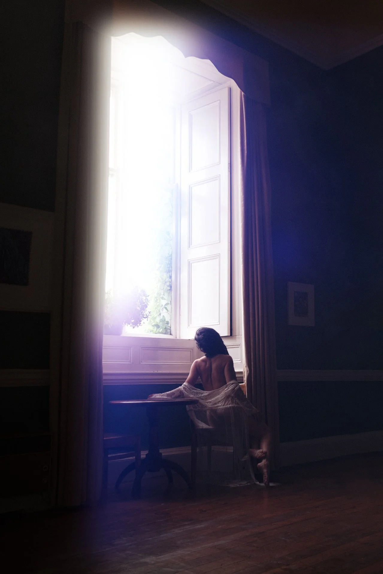 Fine art nude figure seated in soft window light, sheer fabric draped over the body, creating a contemplative silhouette against bright shutters in a dark, moody interior.