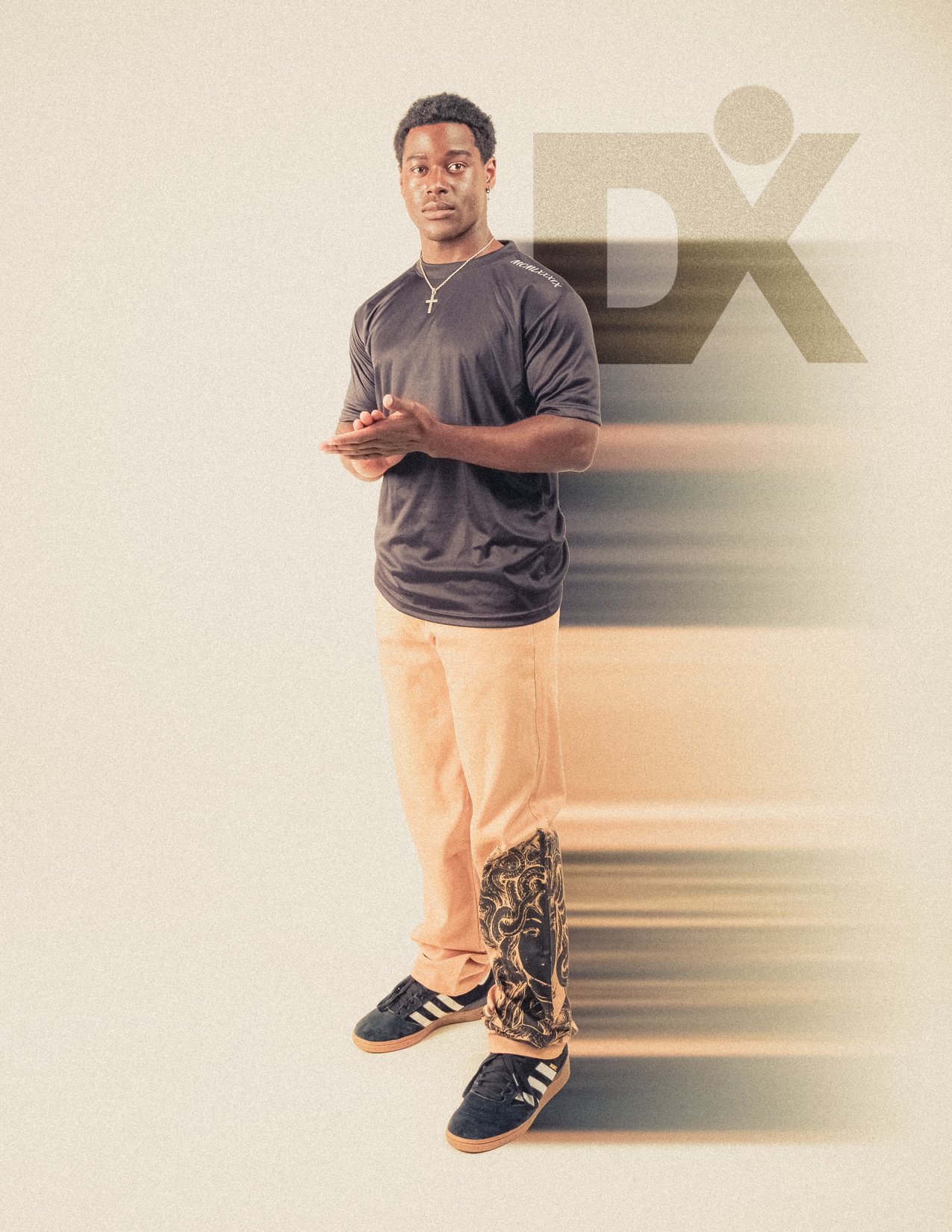 A young man standing against a backdrop with a large 'DX' logo, wearing a black athletic shirt, beige pants with a decorative black and gold design on one leg, black sneakers with white stripes, a kind of jewelry around his neck, and looking directly