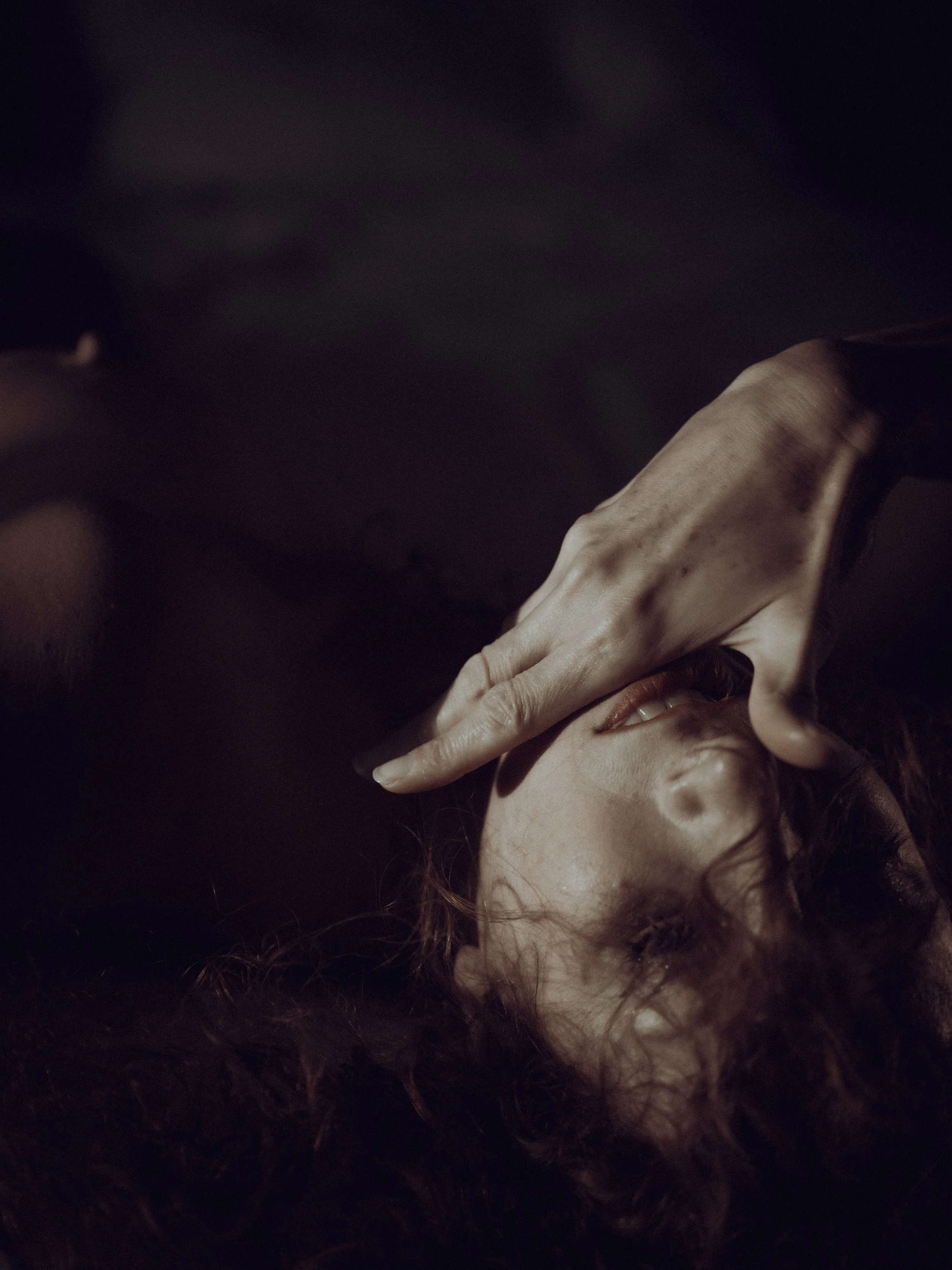 A woman with wavy hair lies on a dark surface with her face partially covered by her hand.