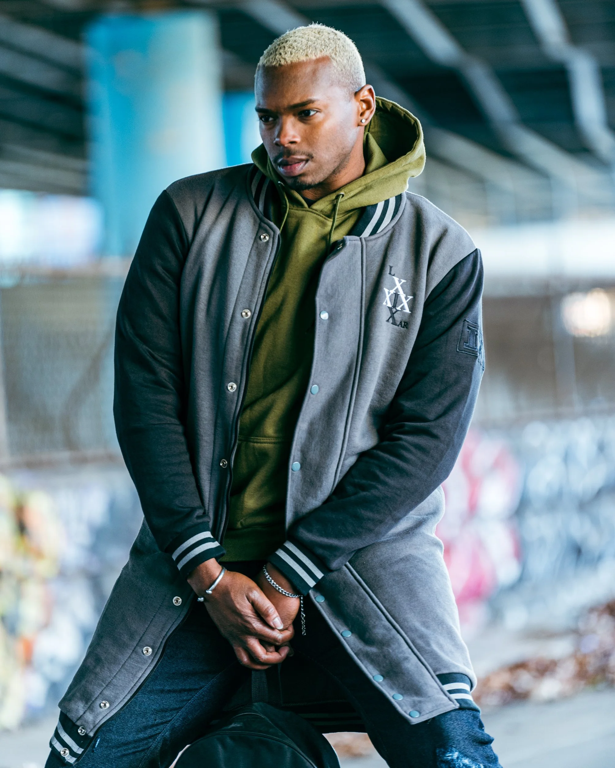 A young man with platinum blonde hair wearing a green hoodie and a gray and black varsity jacket, sitting outdoors under a bridge, holding his hands together in front of him.