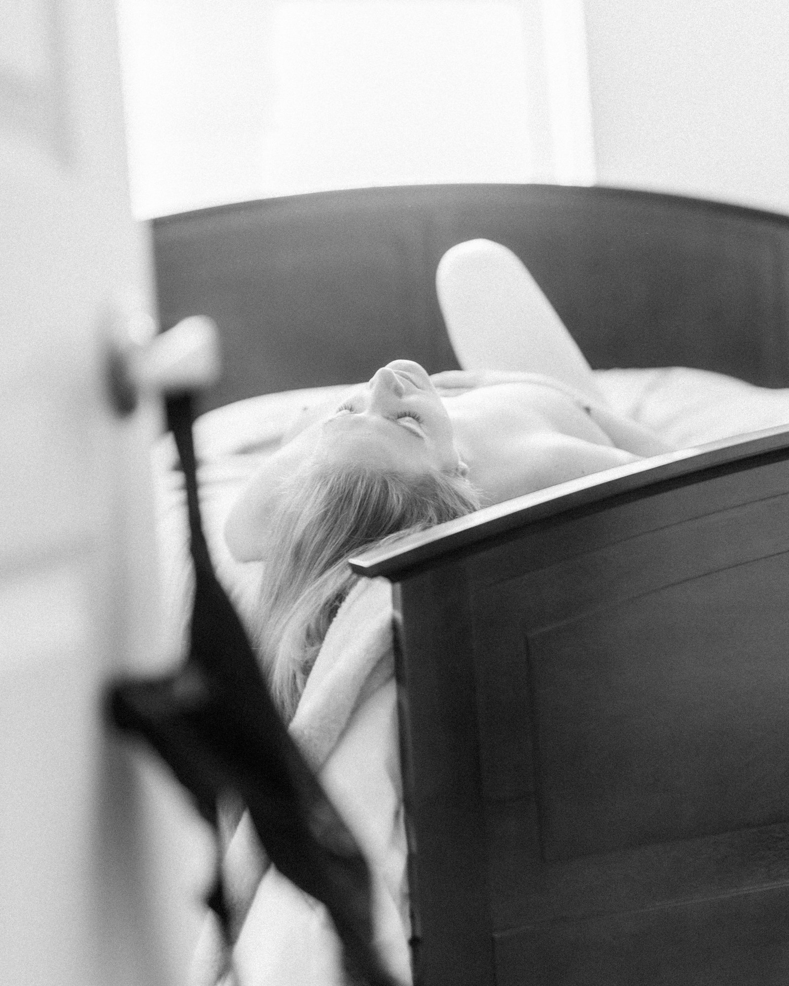 A woman lying in bed, seen through a partially open door in a black and white photograph.
