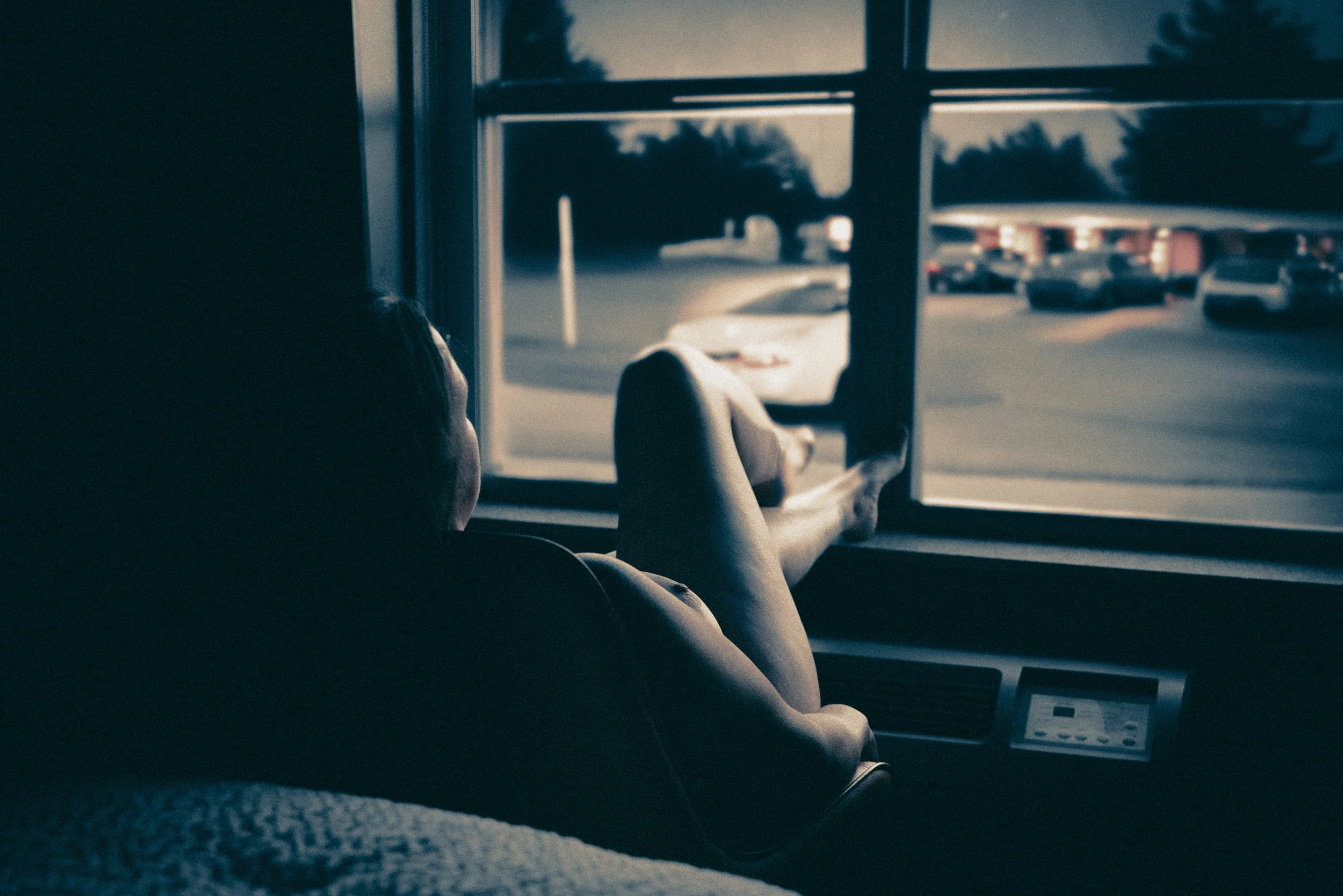 Person resting on a bus seat, looking out the window at parked cars during dusk or dawn.