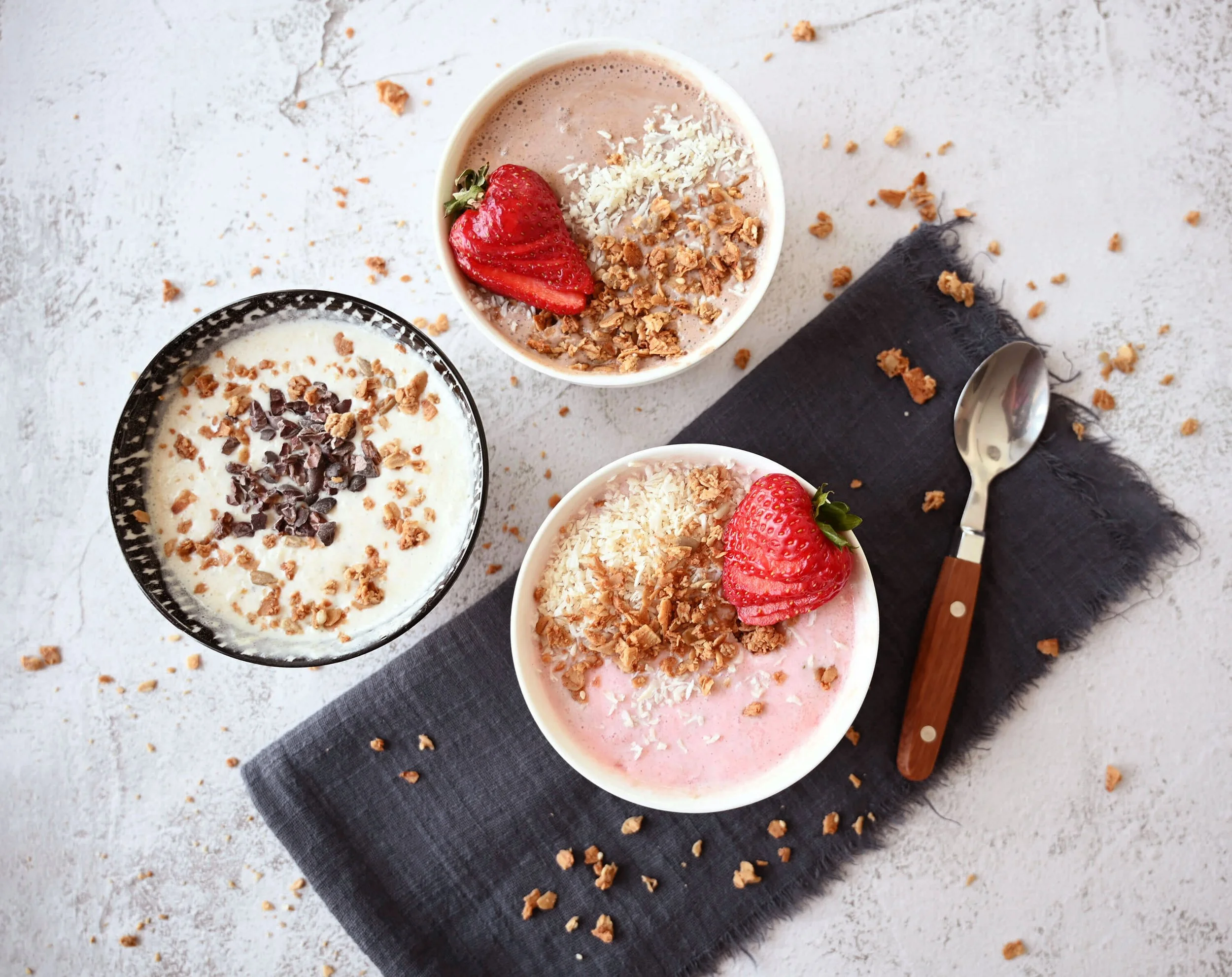 Three bowls of yogurt with strawberries, granola, and chocolate chips on a gray napkin, with a spoon nearby.