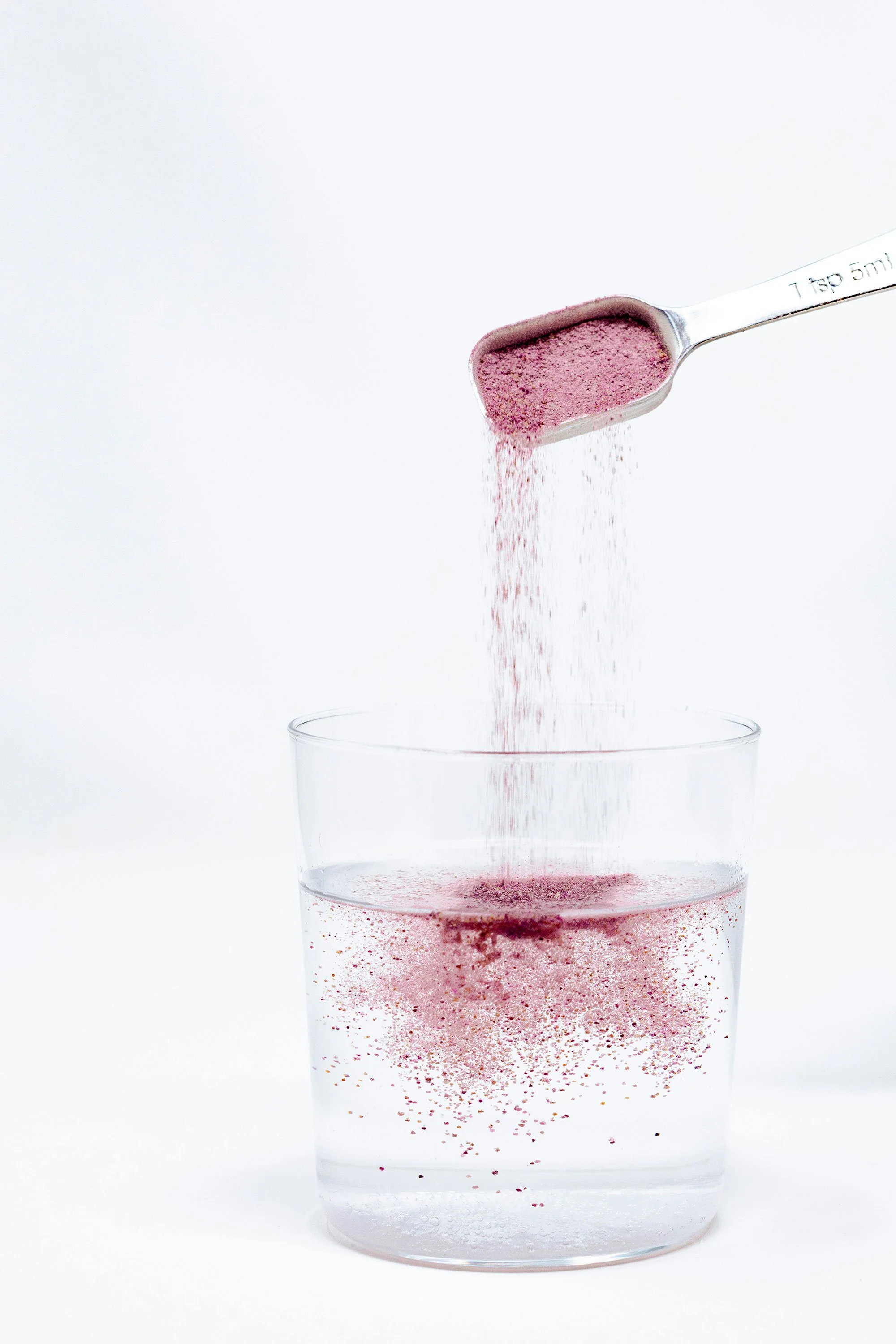 A spoon with pink powder being poured into a clear glass of water.