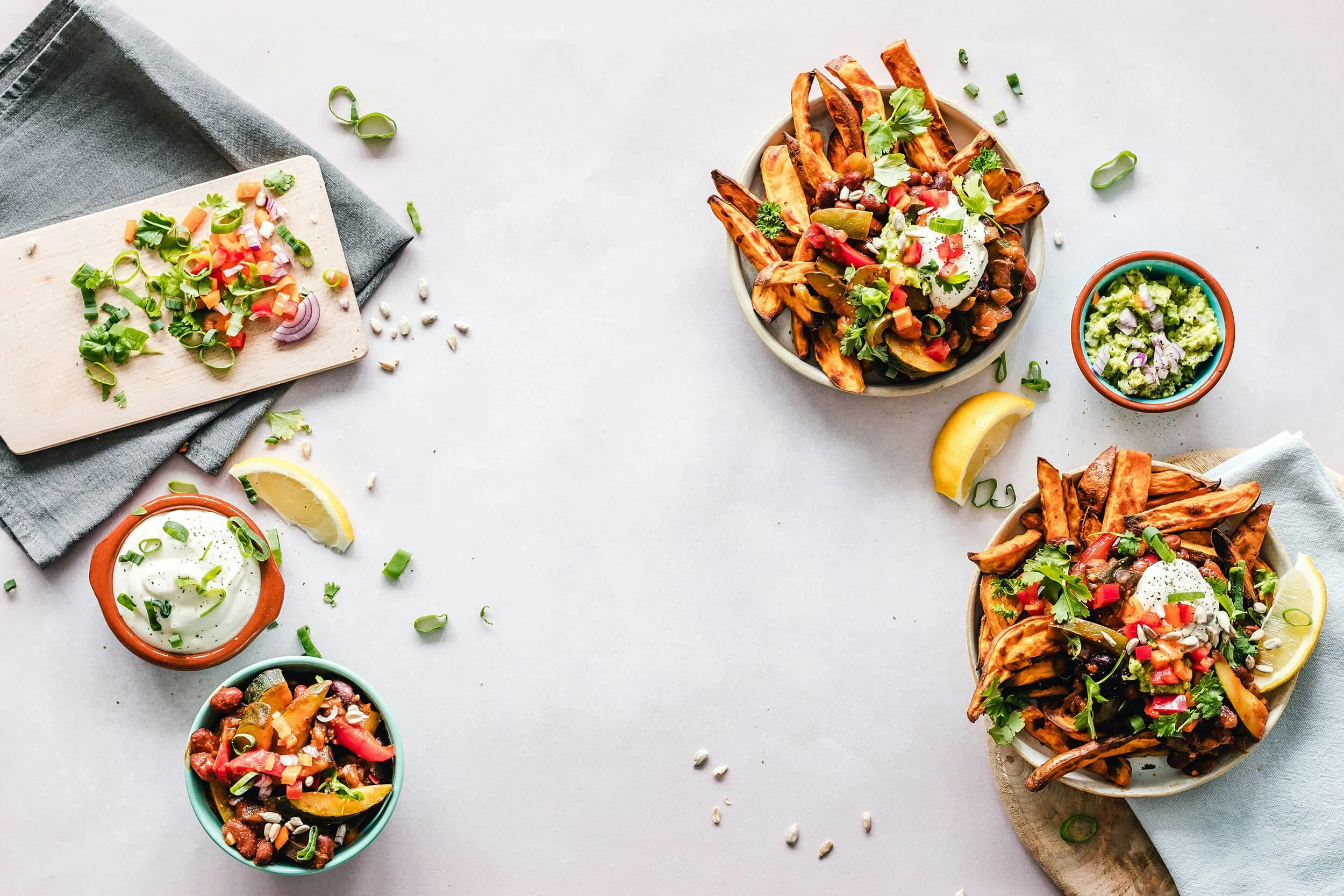 Three plates of loaded sweet potato fries with toppings like sour cream, chopped tomatoes, green onions, and cilantro, along with bowls of guacamole and sour cream, lemon wedges, and scattered green onions and sunflower seeds on a light background.