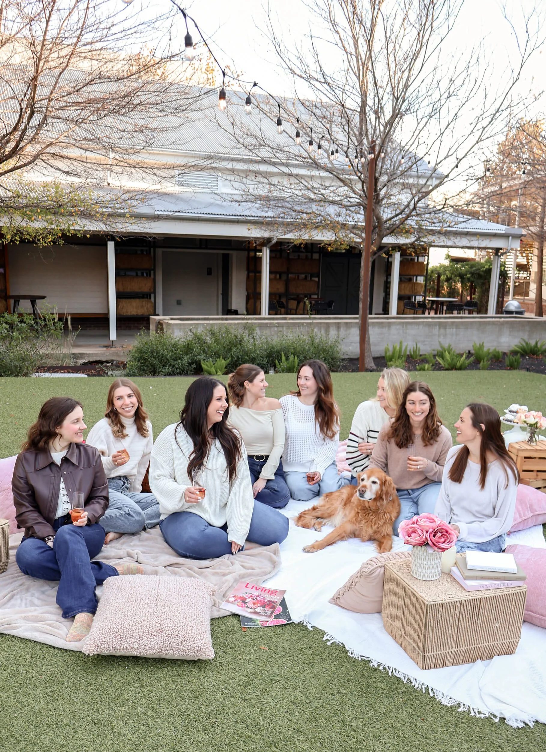 A group of women and a dog having a picnic outdoors on a blanket with pillows, books, and flowers, smiling and talking.