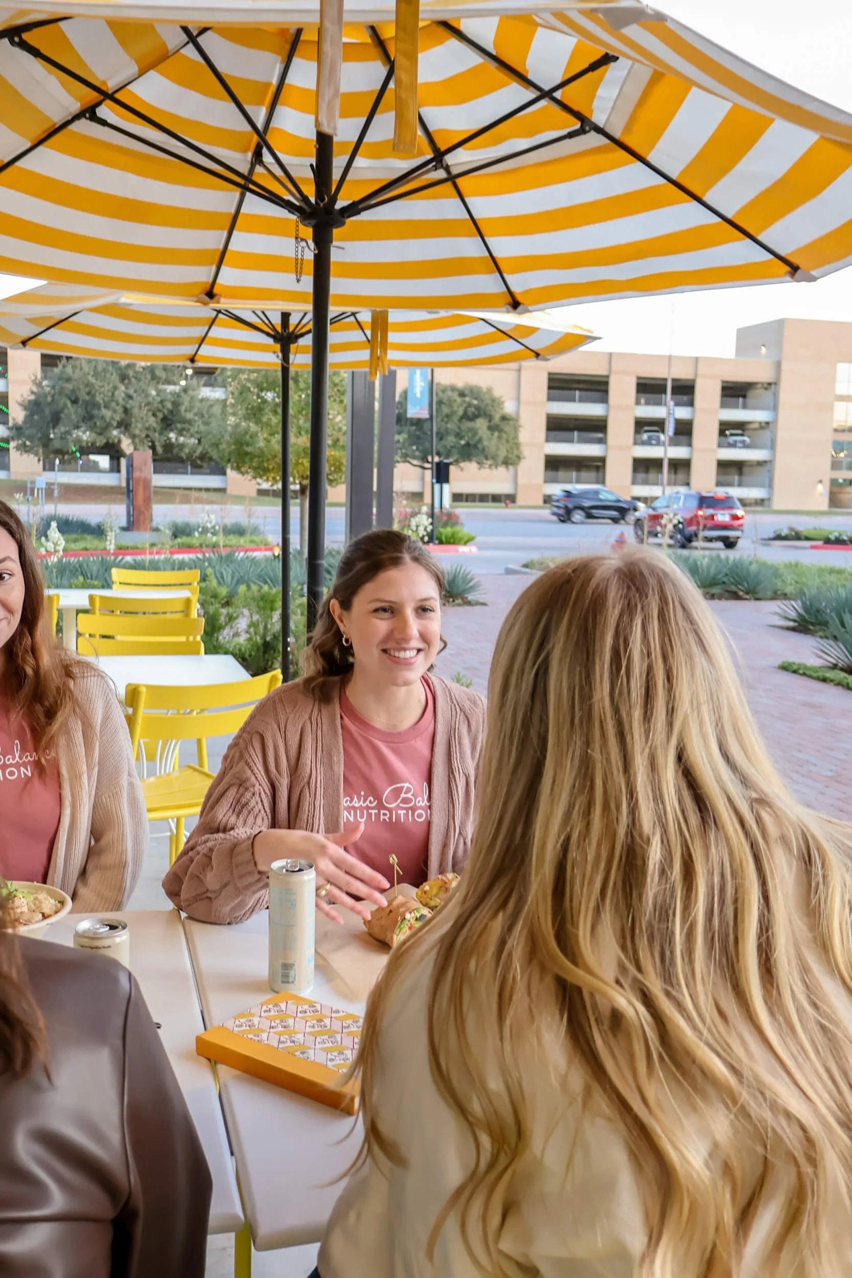 Group of women sitting at a table outdoors, smiling and chatting, with umbrellas overhead and a city street in the background.