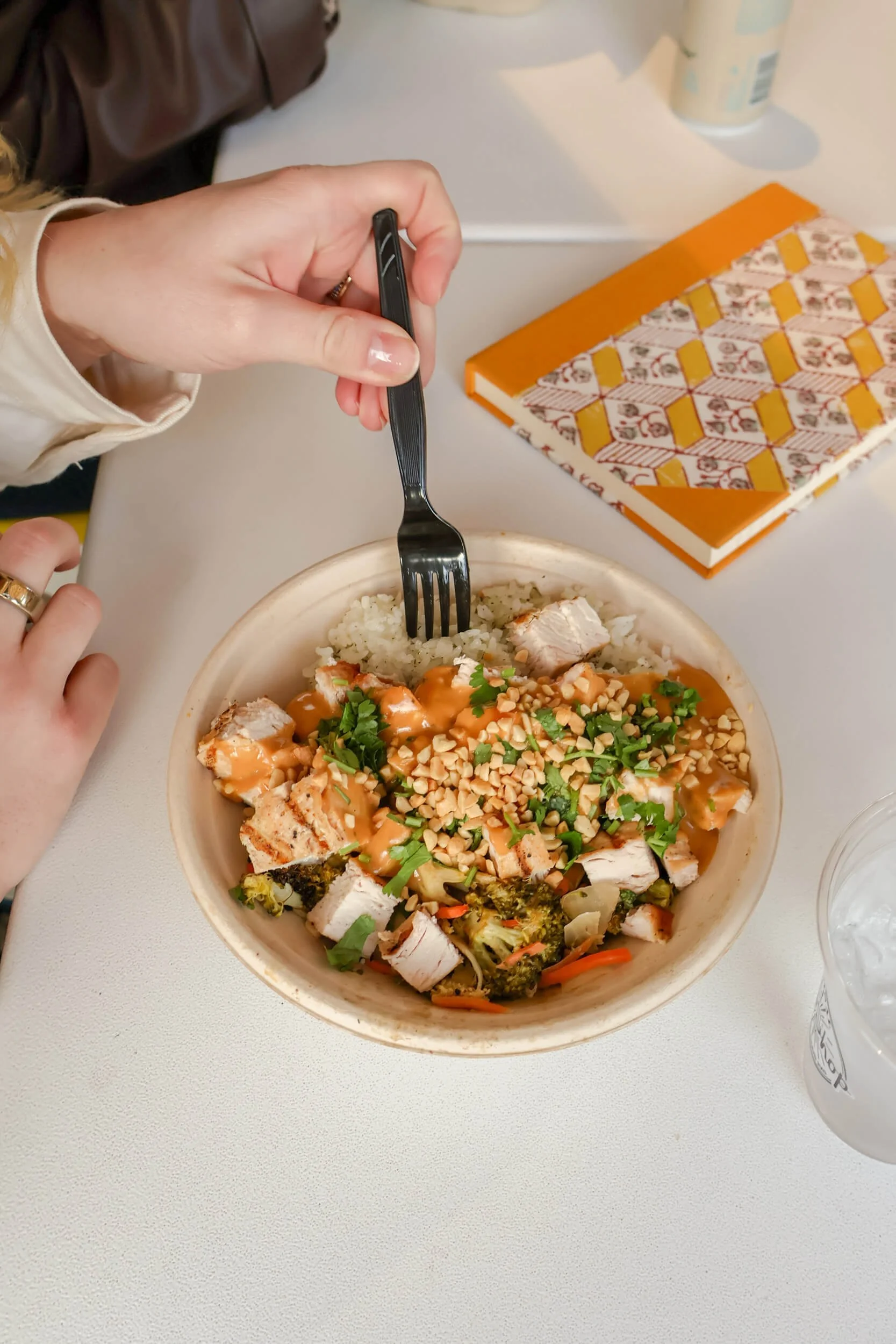 A person holding a fork and about to eat a bowl of rice topped with grilled chicken, peanuts, cilantro, and sauce on a white table with a notebook, a drink, and a bottle in the background.