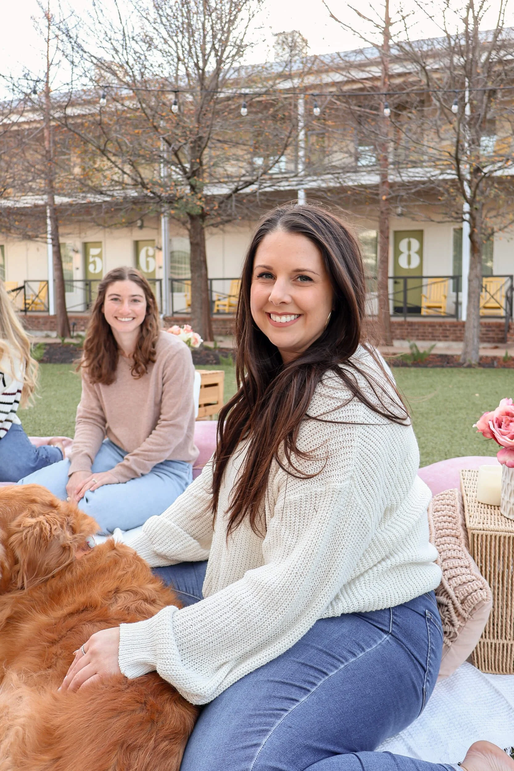 People enjoying a daytime outdoor gathering, sitting on a pink outdoor rug with a golden retriever, with a building featuring numbered doors and leafless trees in the background.