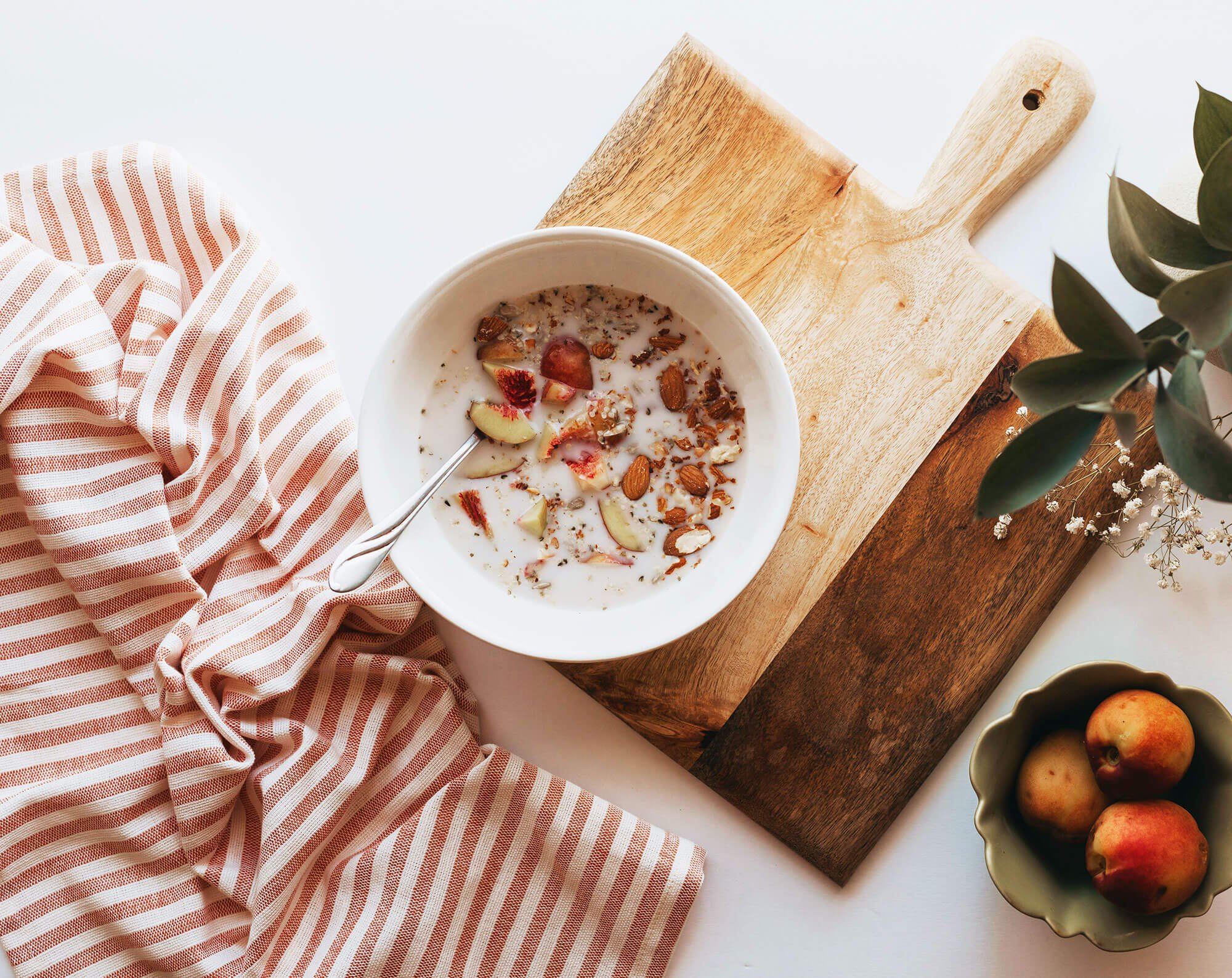 A bowl of cereal with sliced peaches and almonds on a wooden cutting board, with a pink striped cloth to the side and a small bowl of peaches nearby.