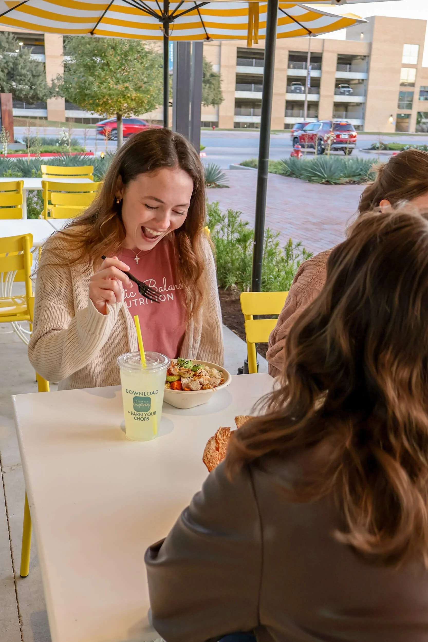 A young woman with long brown hair, wearing a beige cardigan over a pink shirt, is sitting at an outdoor table under a yellow and white striped umbrella. She is happily laughing while holding a fork above a bowl of salad. A drink with a yellow straw and a label that says 'DOWNLOAD EARN YOUR CHOPS' is on the table. Other people are partially visible, and there is a parking lot with cars and a building in the background.