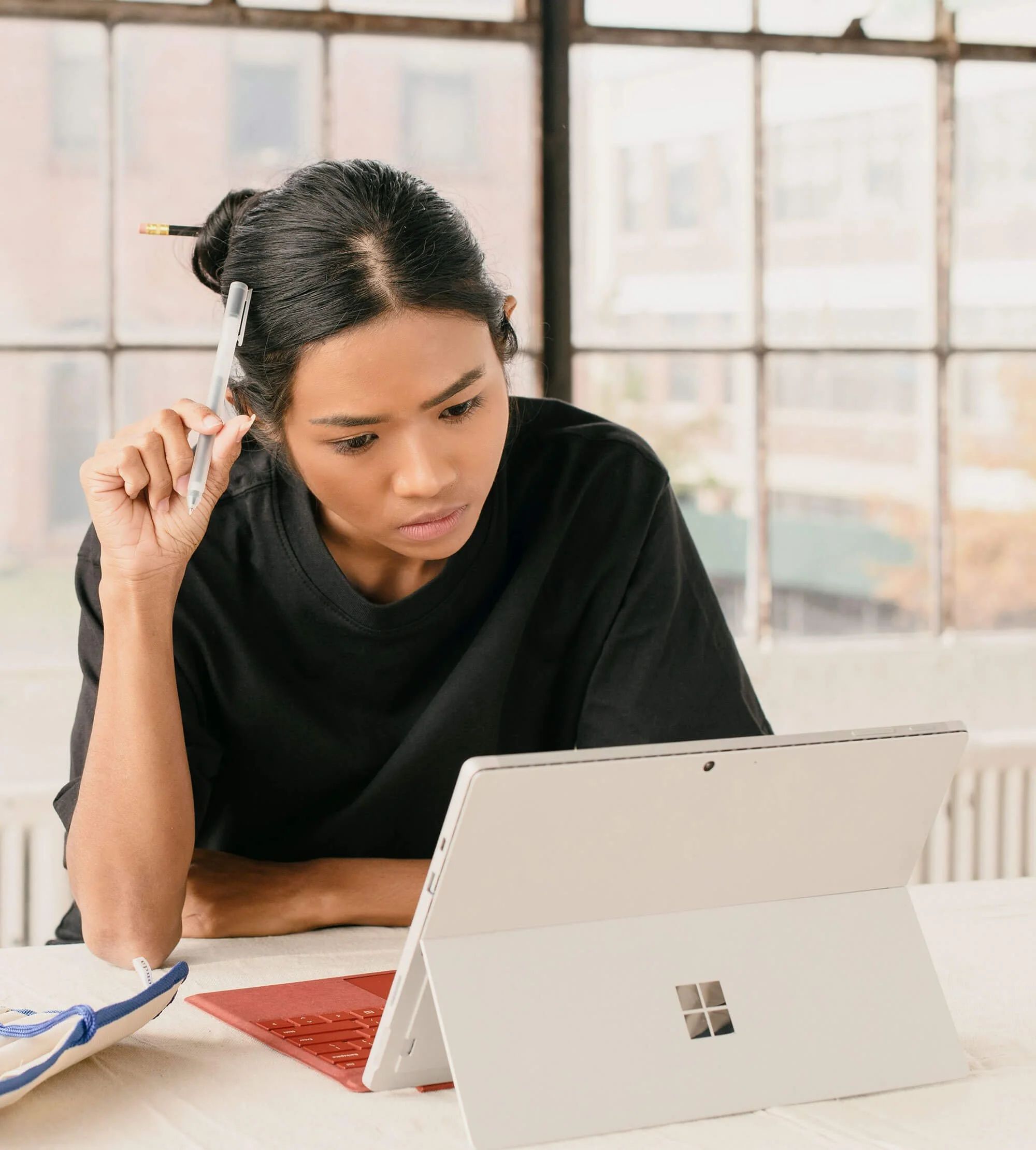 A woman with dark hair tied up, holding a pen to her temple while looking confused at a white Surface tablet on a desk.
