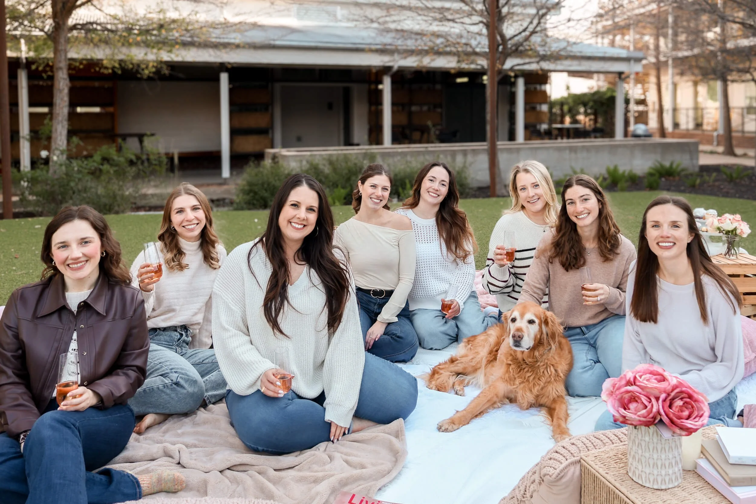 A group of nine women and one dog sitting on blankets, smiling at an outdoor gathering, with drinks in hand and a garden in the background.