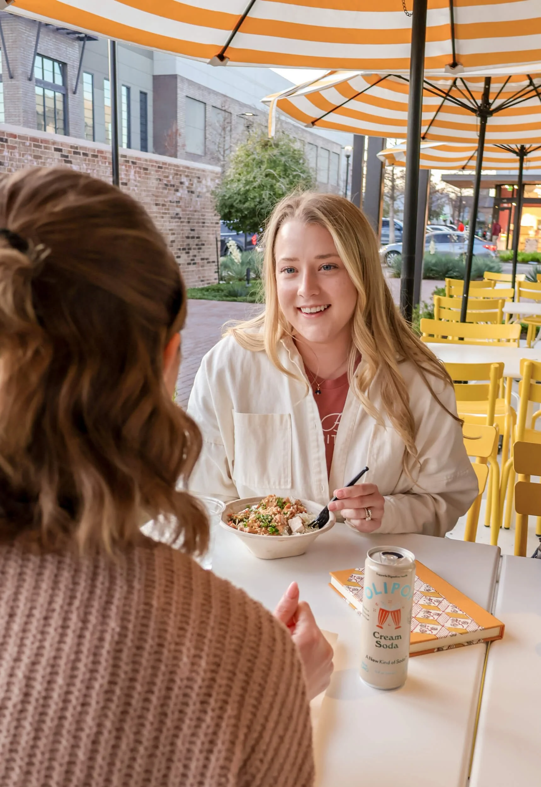 Two women having a meal outdoors at a restaurant. One woman is smiling and holding a fork, while the other woman, with brown hair, is seen from behind. There is a bowl of food and a can of cream soda on the table.
