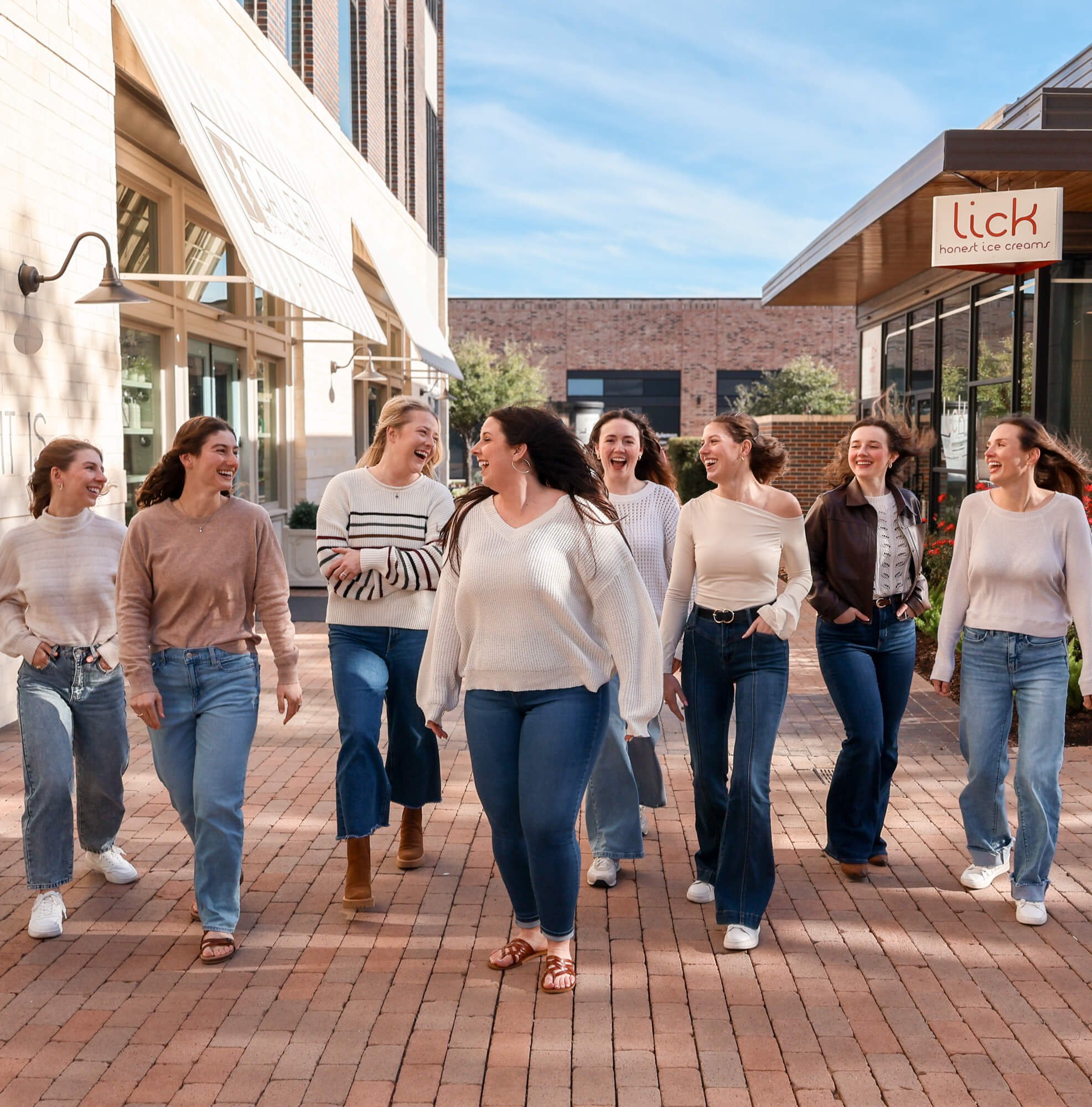 Eight women walking and laughing outdoors on a brick-paved street near shops and cafes.