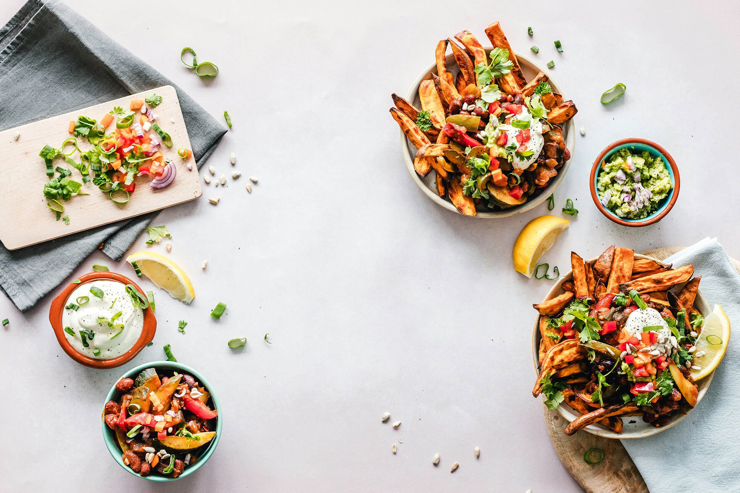 Three bowls of loaded sweet potato fries topped with vegetables, herbs, and sauces, along with a small bowl of guacamole, sour cream, and chopped green onions on the side, on a white surface with lemon wedges.