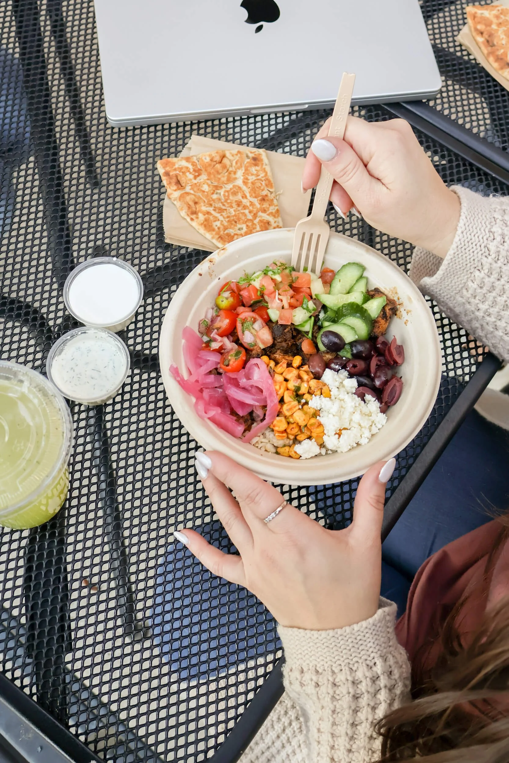 Person holding a bowl of Mexican food with tomatoes, cucumbers, pickled onions, corn, cheese, and black olives, at an outdoor table with a laptop, pizza slice, drinks, and utensils.