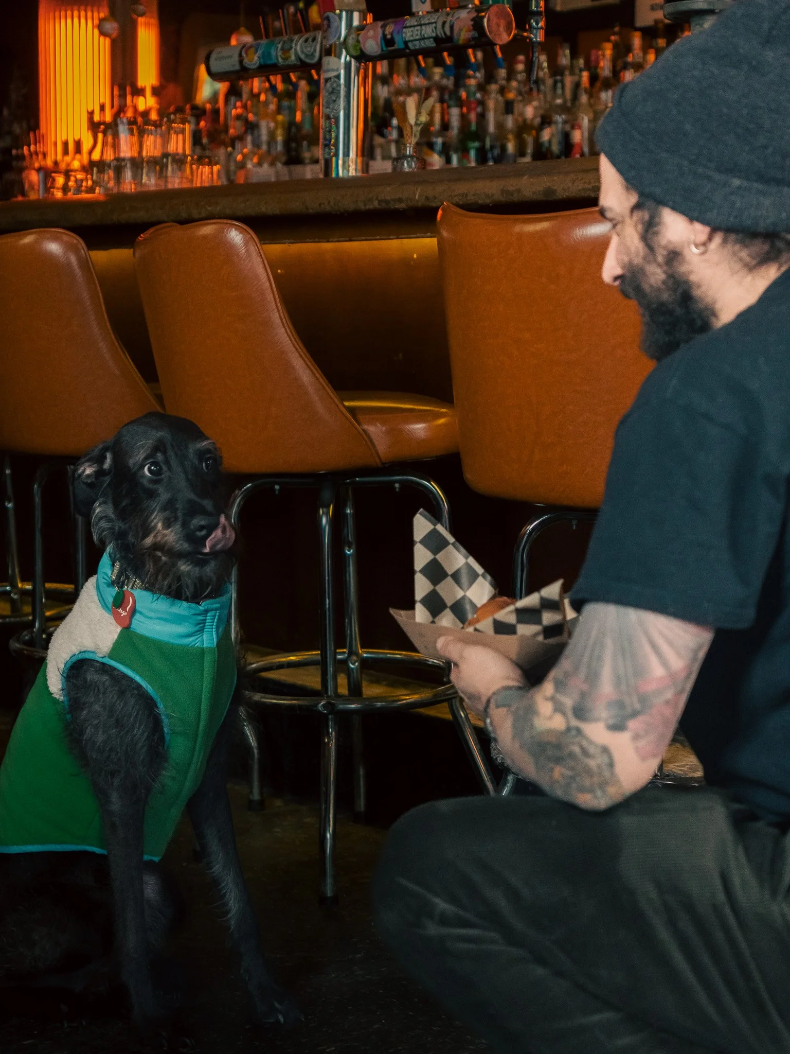 A man with tattoos on his arm, wearing a beanie and black t-shirt, kneels near a black dog wearing a green coat inside a bar. The man is holding a tray of food, and the dog is sitting on the floor looking up at the man.