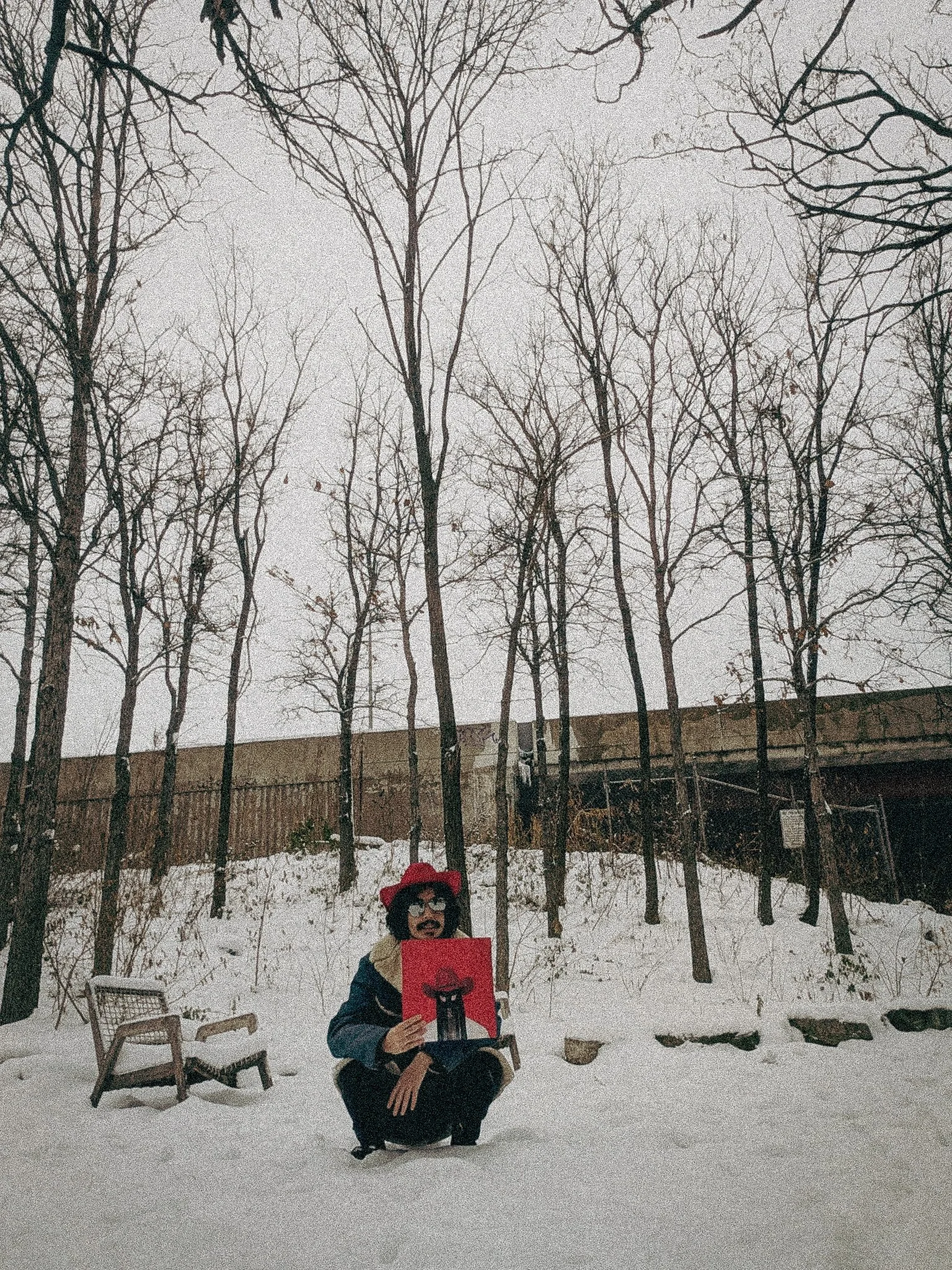 Person in a red hat and sunglasses squatting in snow, holding a red painting of a black cat with a red hat, with a broken chair nearby, trees in the background, and a fence and bridge behind them.